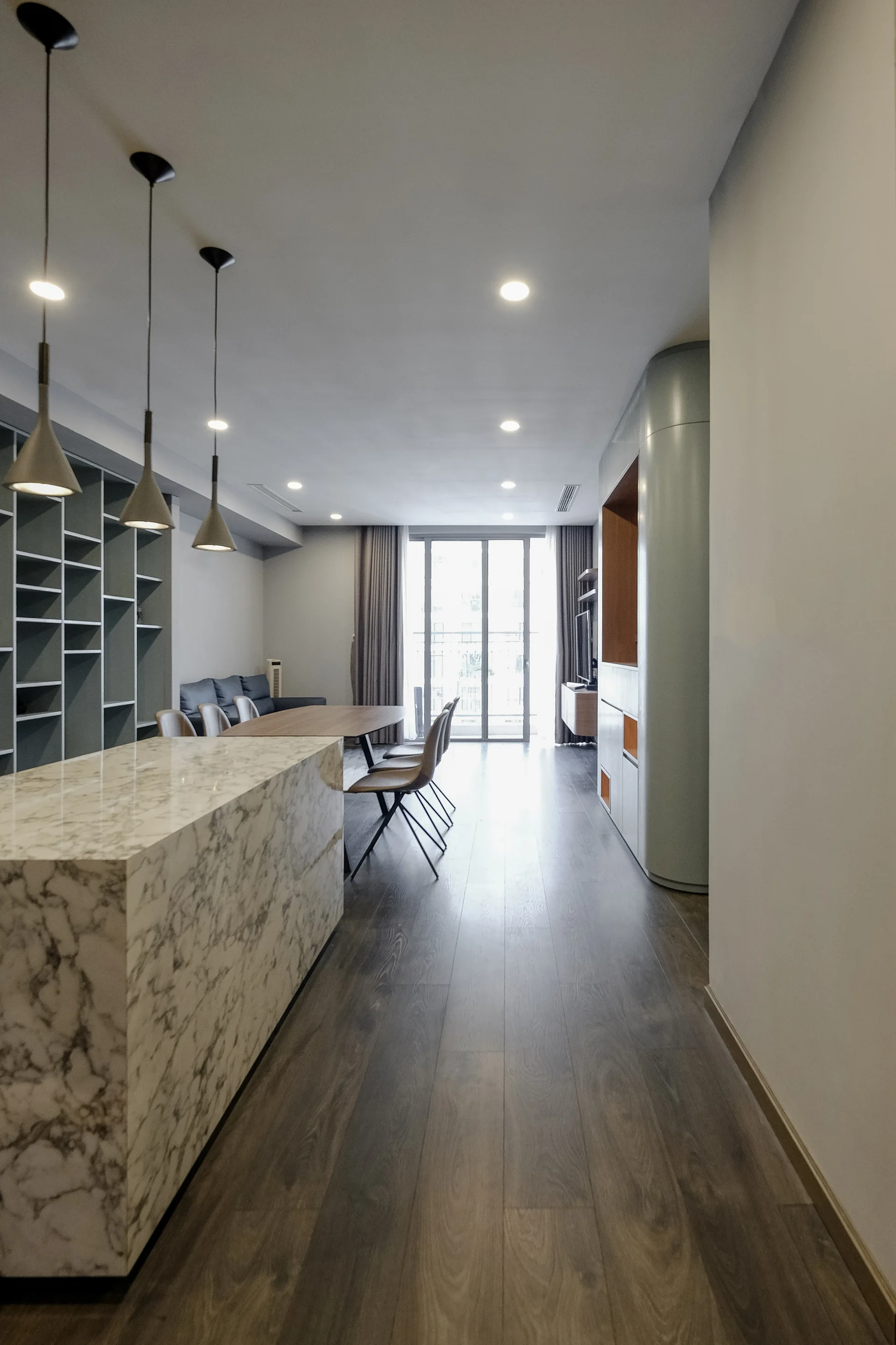 An open-concept apartment interior features a veined marble kitchen island, pendant lights, a dining table, and built-in gray shelving.