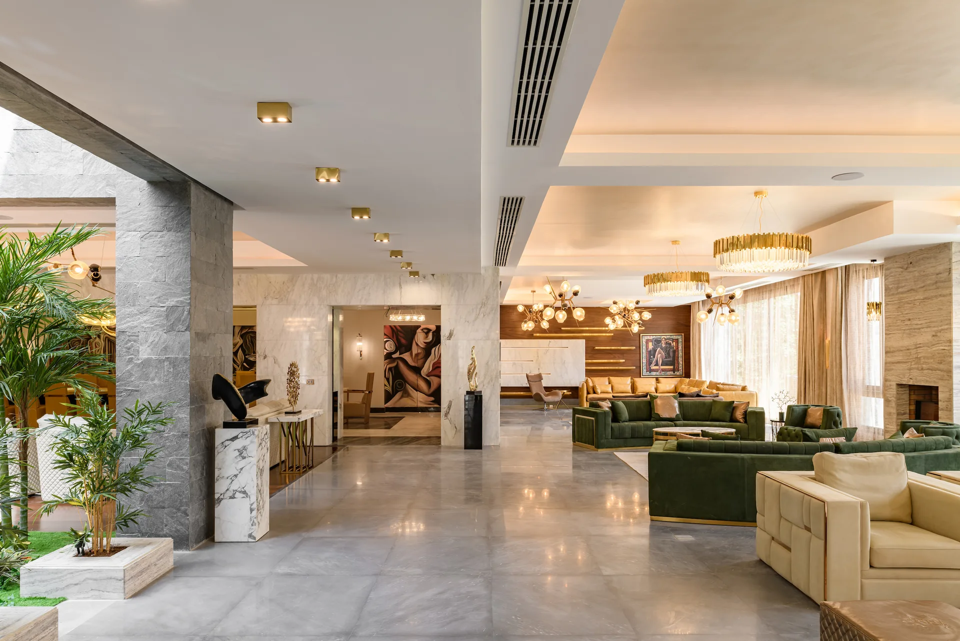 An open-plan living room with green velvet sofas, marble wall panels, polished grey flooring, and glowing gold chandeliers hanging from a tiered ceiling.