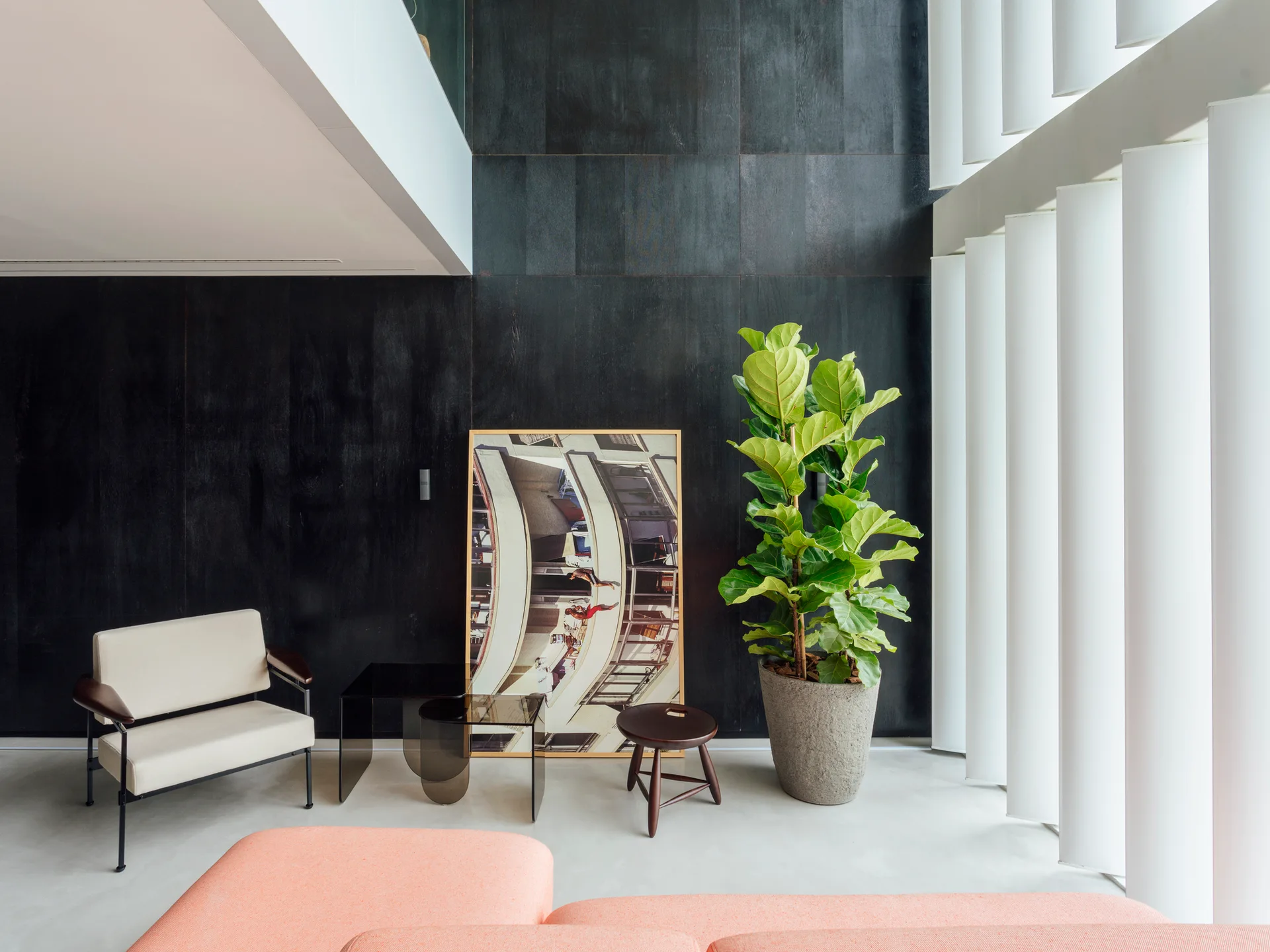 A modern living room featuring black wood wall paneling, a peach sofa, a white armchair, and vertical window louvers next to a potted plant.