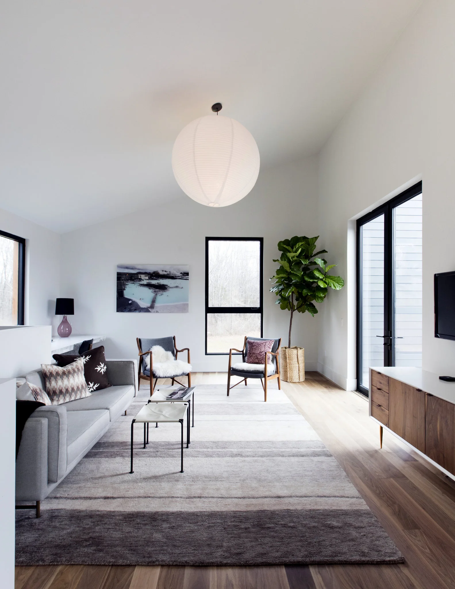 A modern living room features a gray sofa, two wooden armchairs, a large paper globe pendant light, and black-framed windows.
