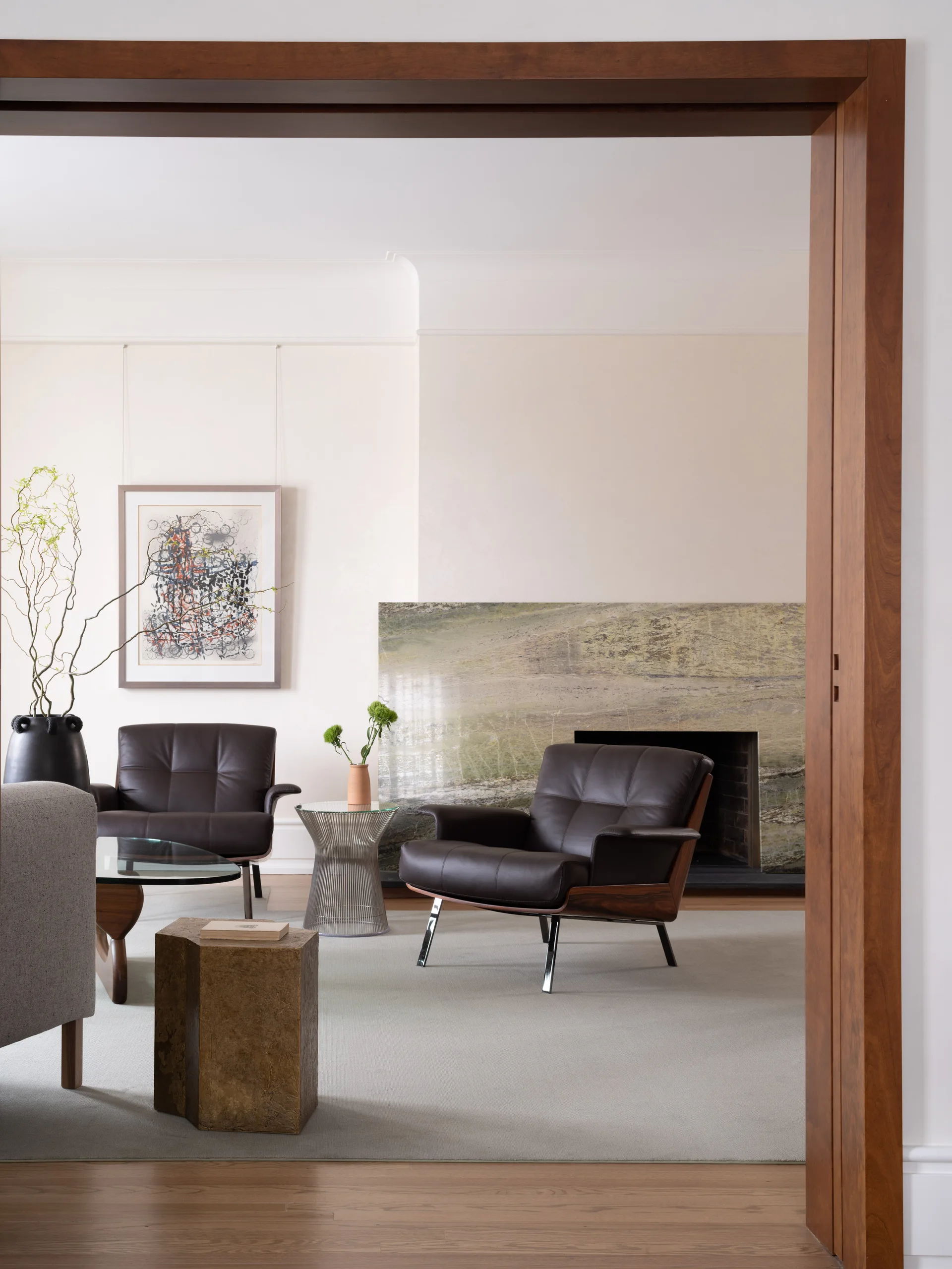 A modern living room viewed through a wood doorway, showcasing two leather armchairs, a large green stone fireplace, and a gray area rug.