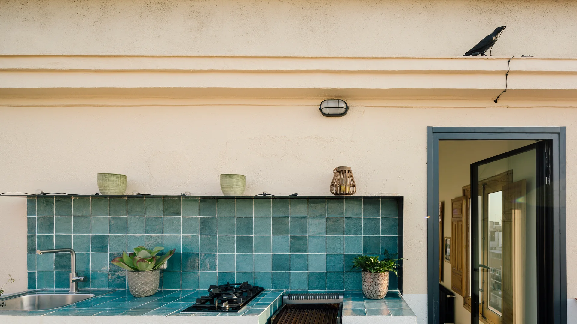 An outdoor kitchen features a glossy teal tiled countertop and backsplash set against a cream stucco wall with an open doorway.