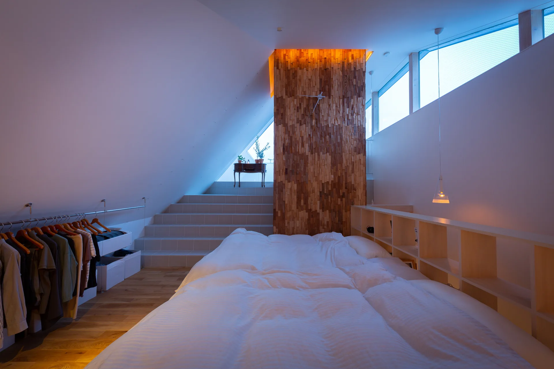 A modern attic bedroom features a low bed with white bedding, a blonde wood headboard shelf, and a textured wooden accent wall near a staircase.