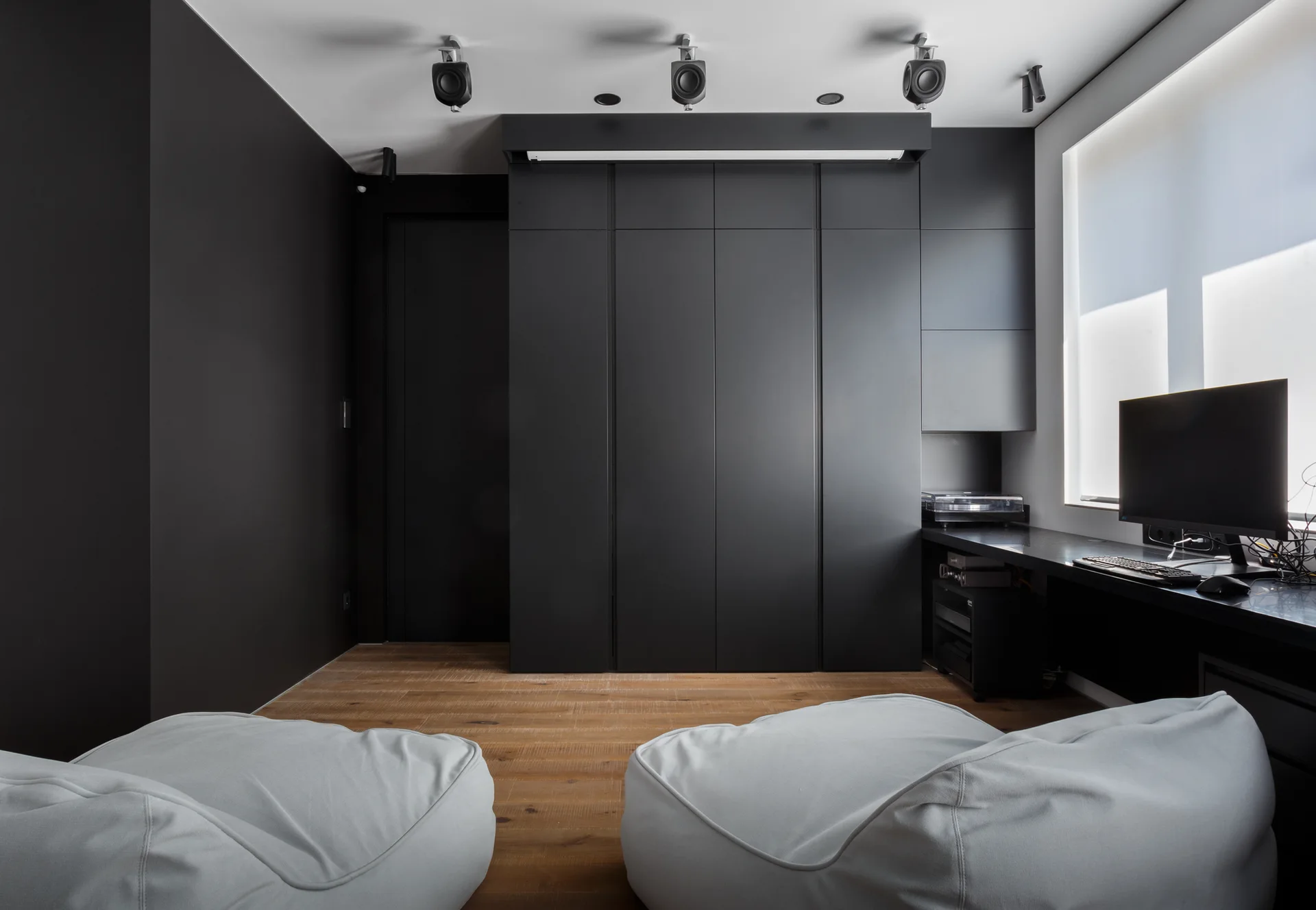 A minimalist home office featuring matte black built-in cabinetry, light gray beanbag chairs on wood floors, and a desk workspace beside a window.