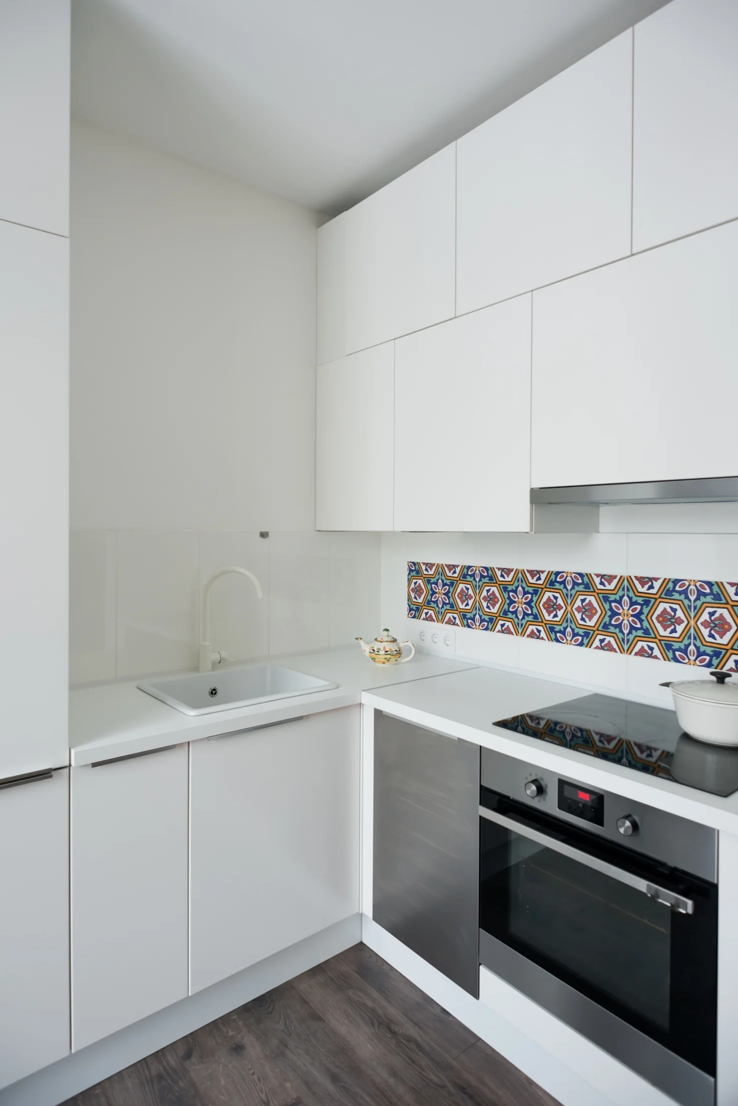A minimalist corner kitchen features white flat-front cabinets, an undermount white sink, and a colorful geometric patterned tile backsplash.