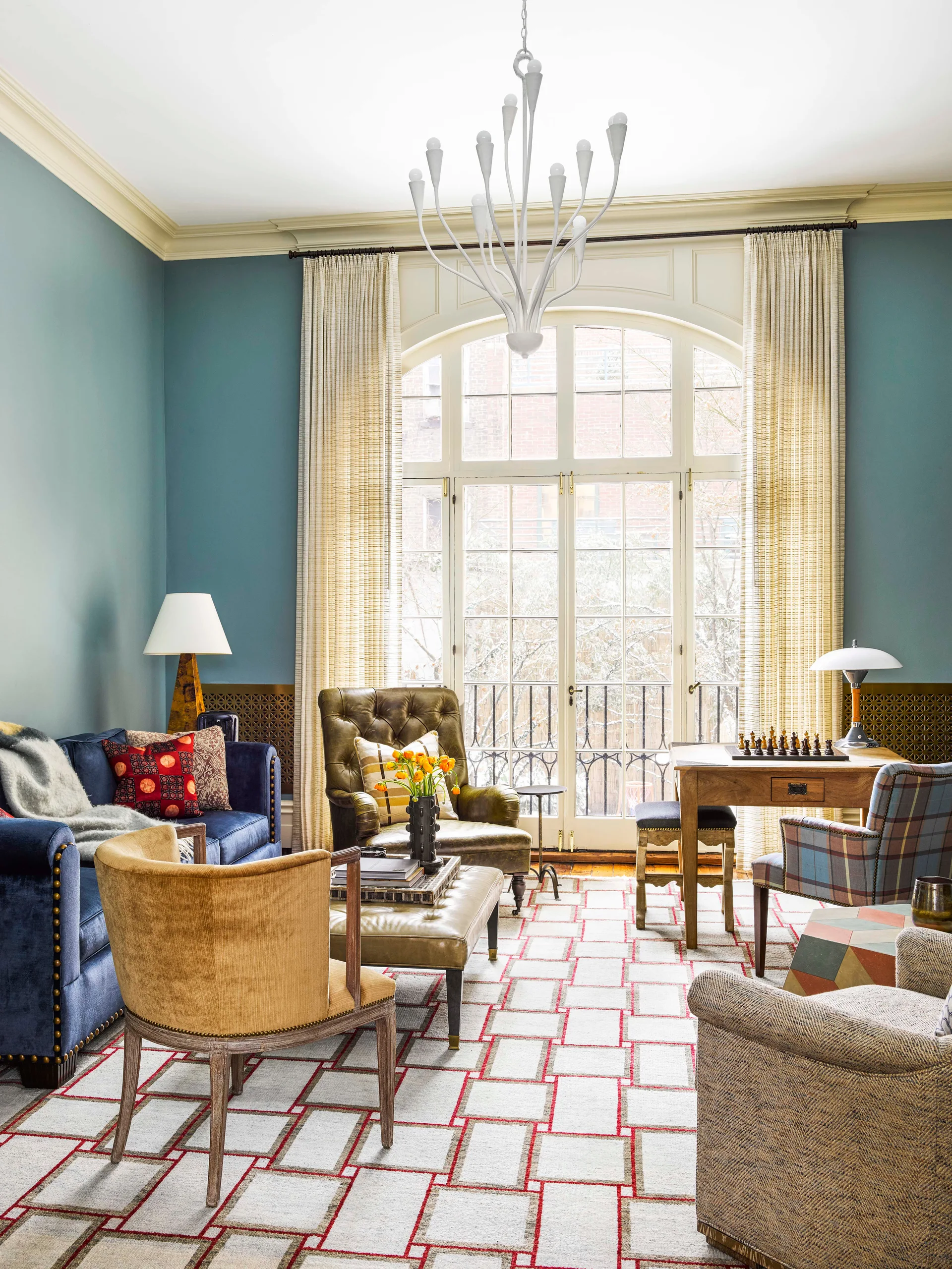 An eclectic living room with blue walls features a large arched window, a blue velvet sofa, a tufted leather armchair, and a wooden chess table.