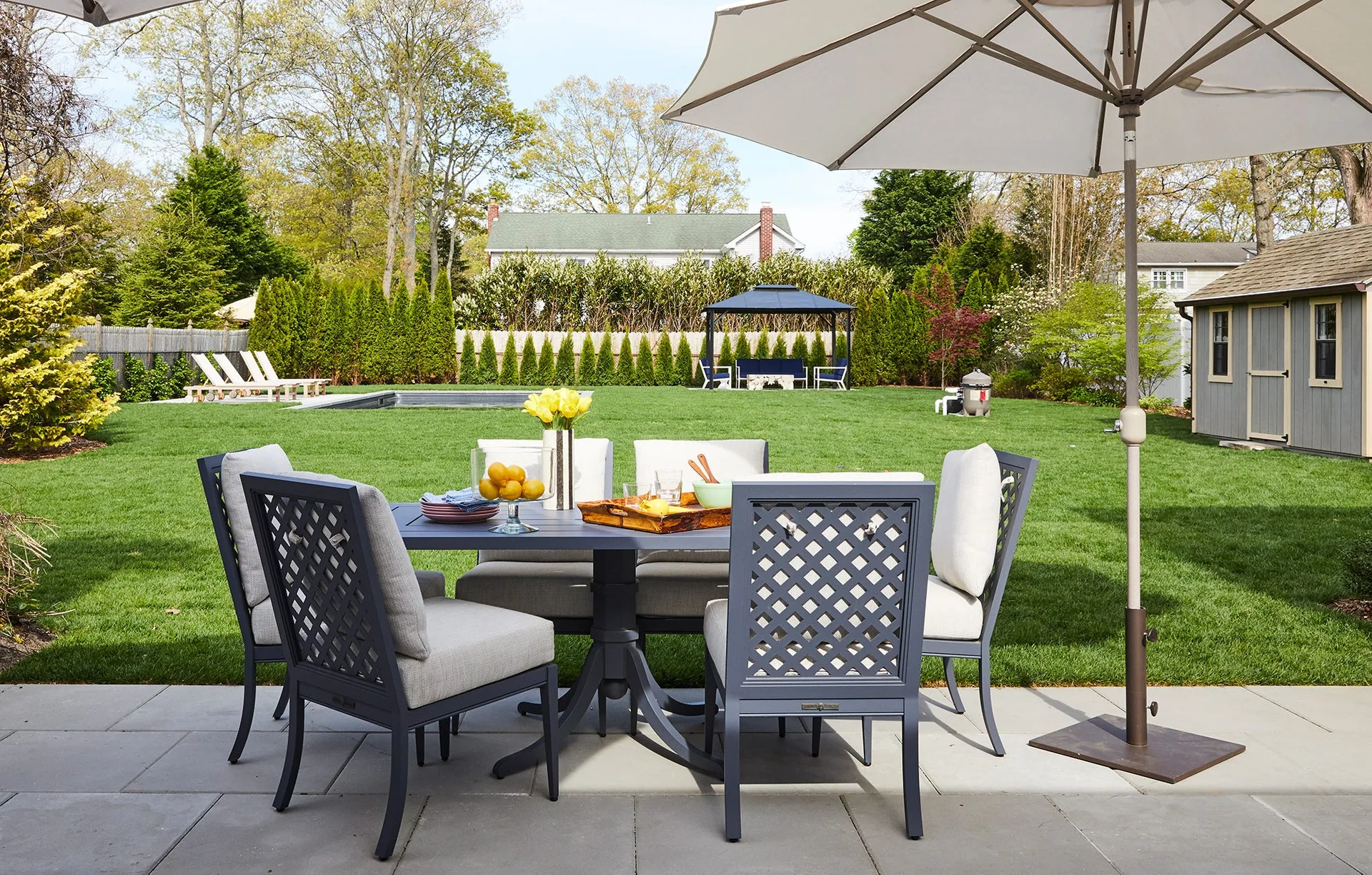 A dark grey metal dining set with neutral beige cushions is arranged on a stone tile patio next to a spacious green lawn.