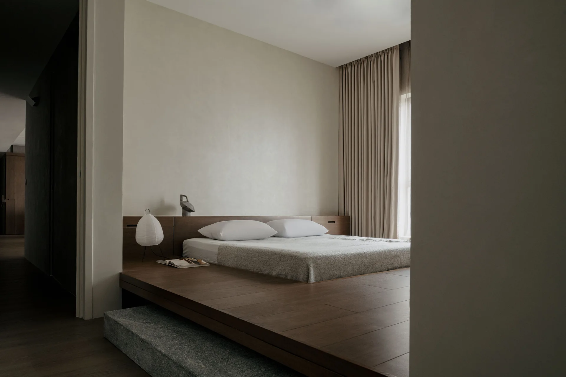 A minimalist bedroom features a low wooden platform bed, textured beige plaster walls, full-length neutral drapes, and a grey stone entry step.