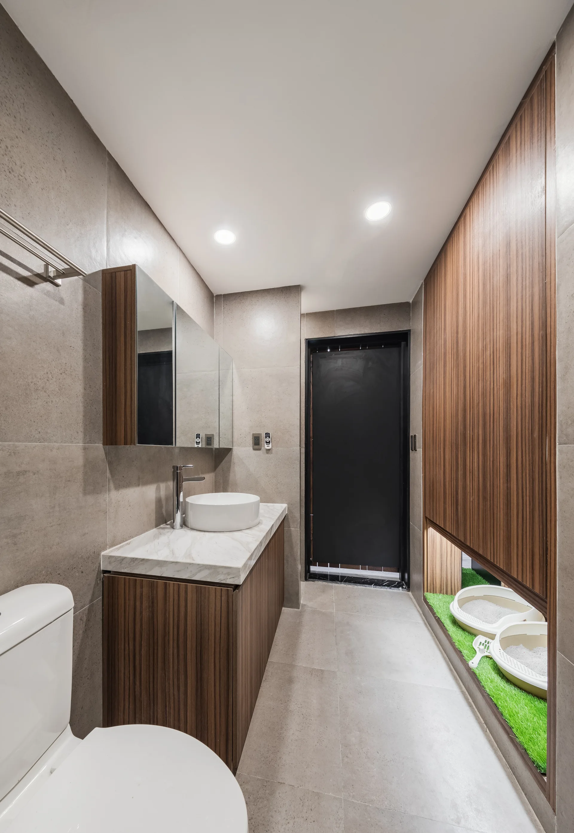 A modern bathroom features grey tile walls, a wood vanity with a white vessel sink, and a built-in floor recess containing cat litter boxes.