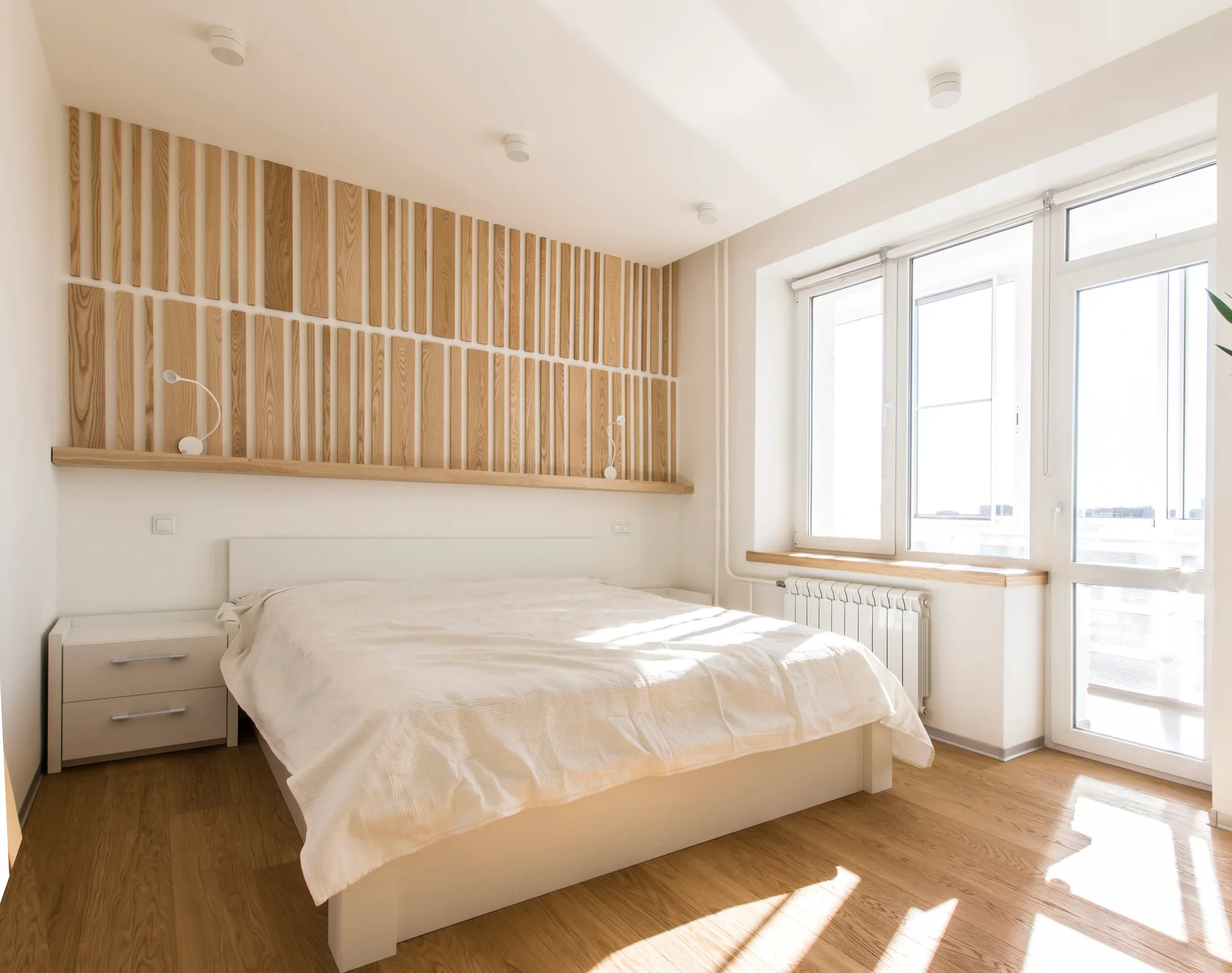 A modern white bedroom features a wood slat accent wall with a matching horizontal ledge above a white bed.