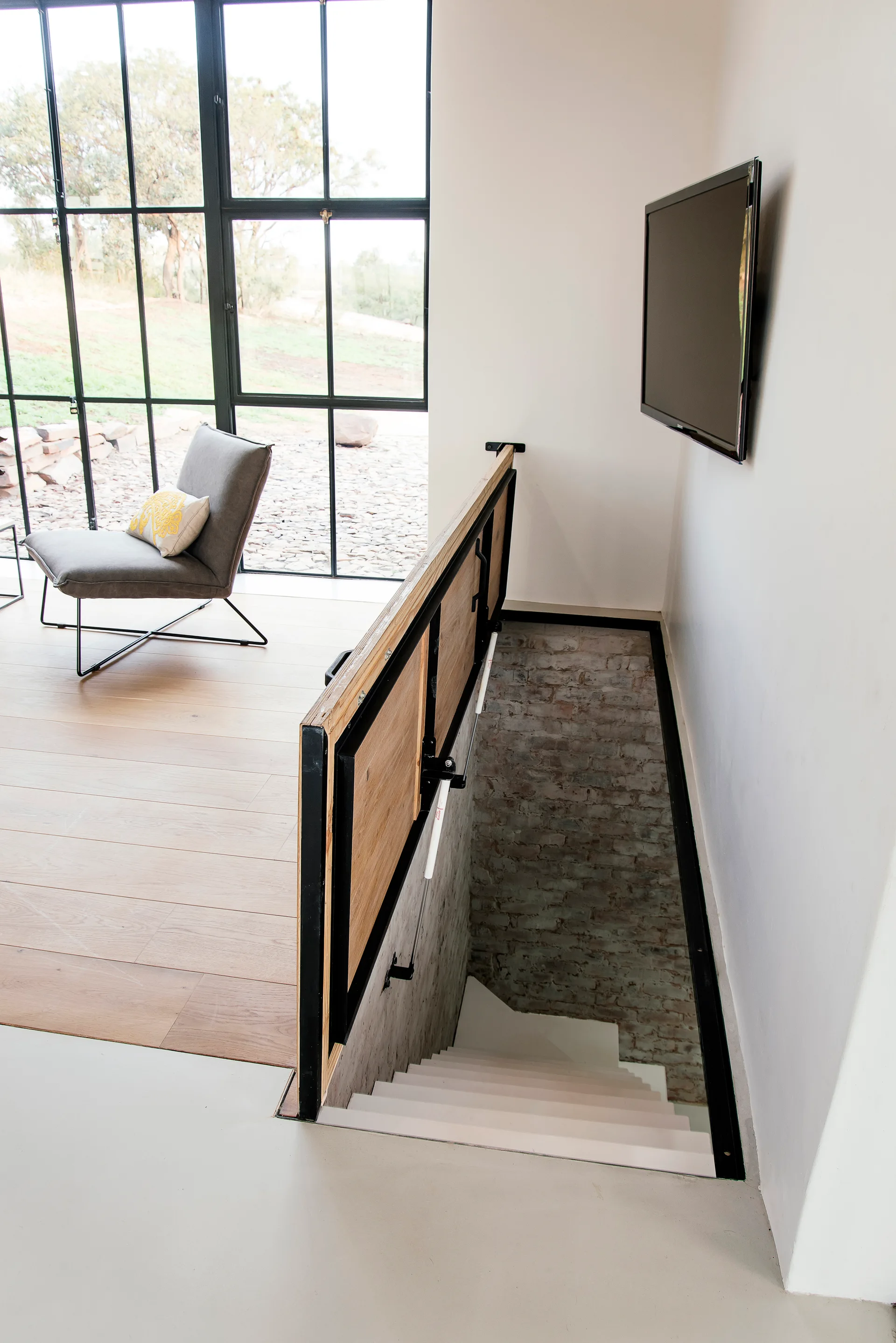A modern staircase with white steps and an industrial wood and metal railing leads down past an exposed brick wall next to a large grid window.