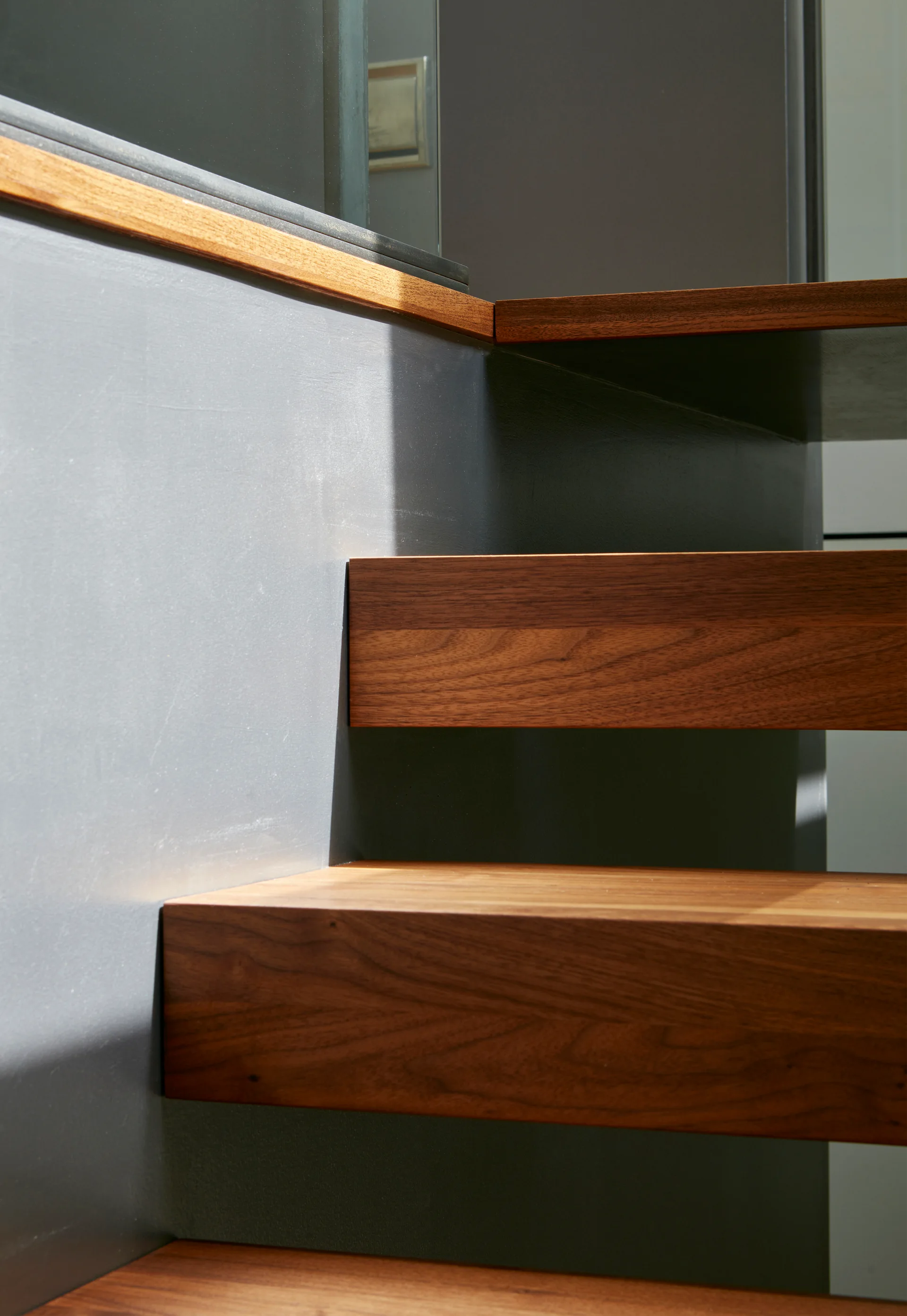 A close-up of floating wood staircase treads mounted against a smooth matte grey wall with natural sunlight casting geometric shadows.