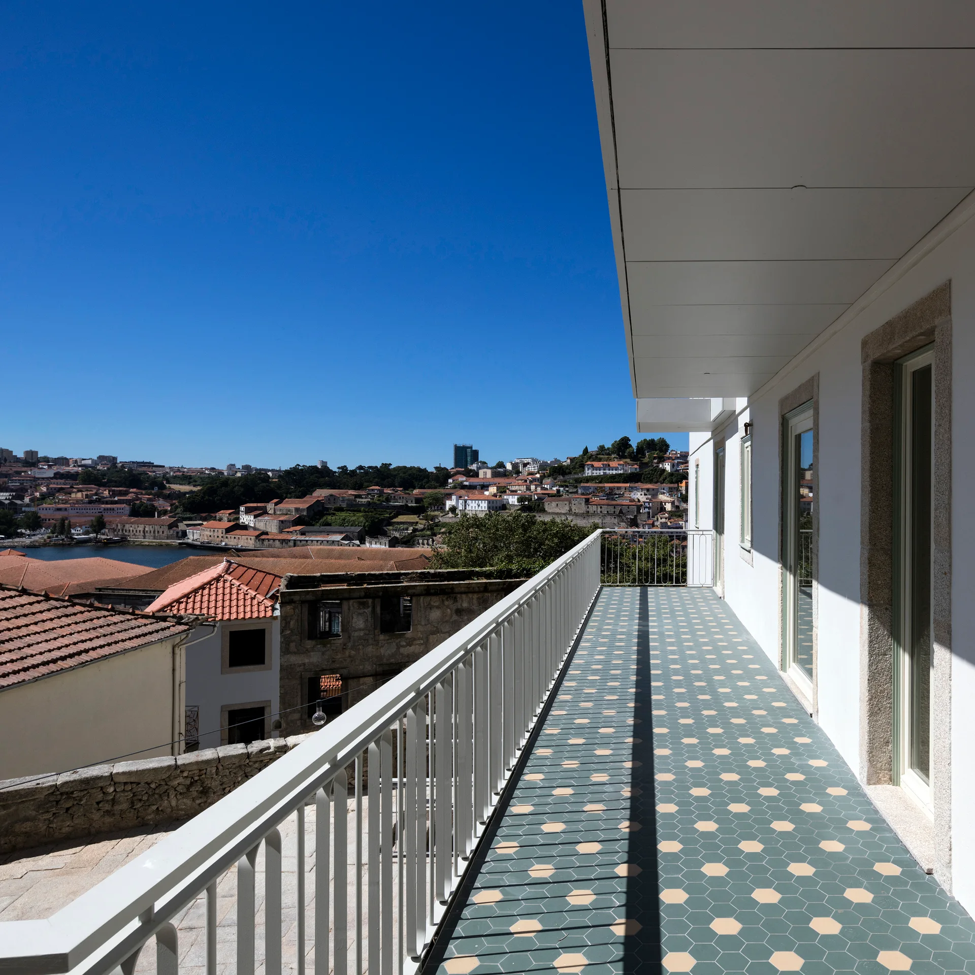 A narrow white balcony featuring blue and beige hexagonal floor tiles and a white metal railing overlooking a historic city under a clear blue sky.