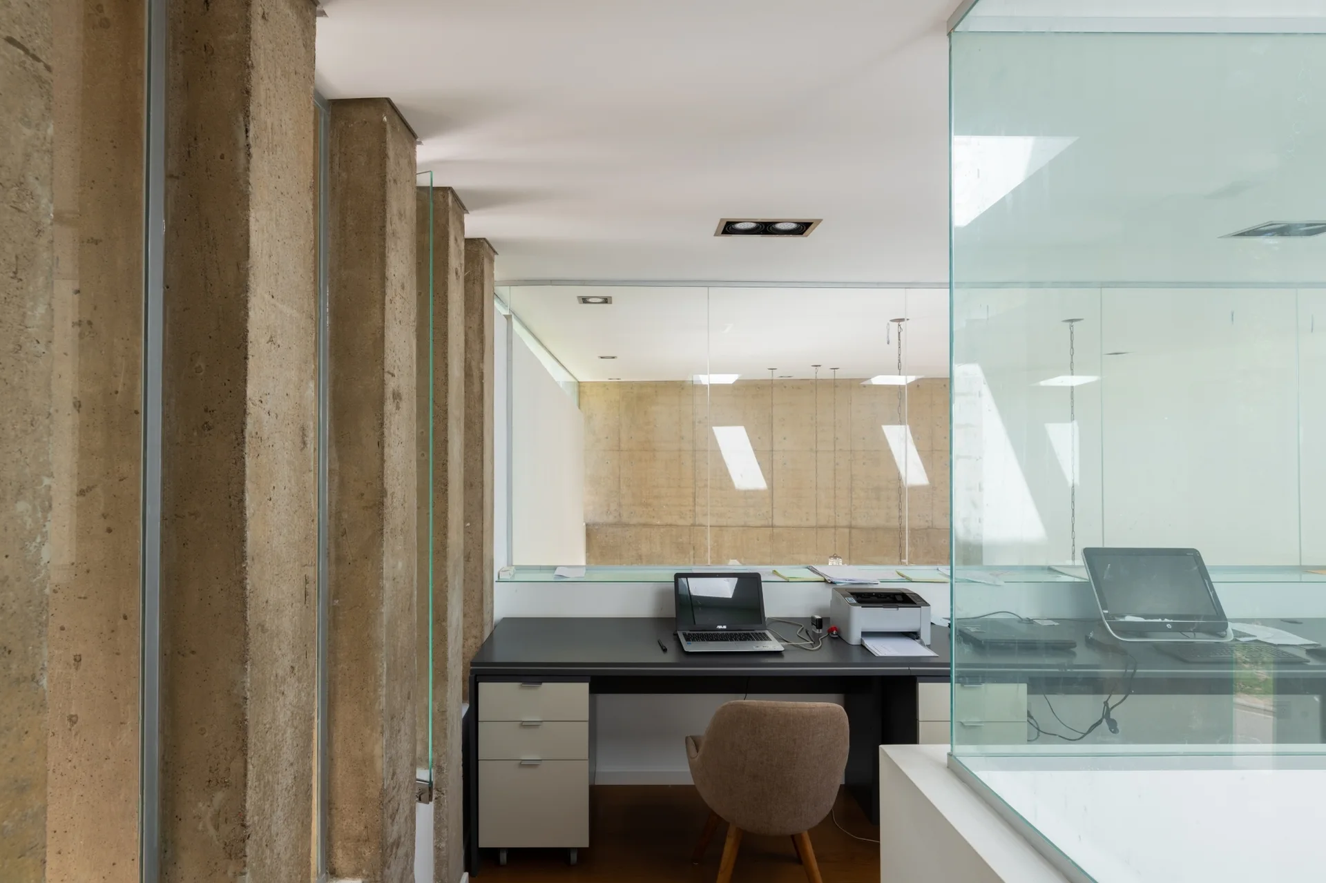 A minimalist home office features a dark desk with a laptop and printer, surrounded by raw concrete columns and glass partitions.