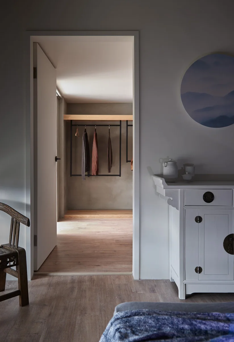 An open doorway connects a room with a white cabinet to a walk-in closet featuring a minimalist black hanging rack and light wood flooring.