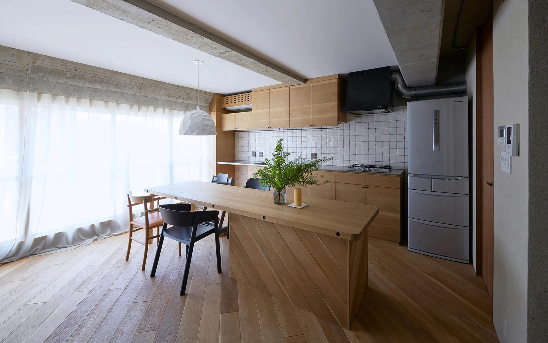 A modern wood kitchen featuring an integrated wooden dining island, a square tile backsplash, an exposed concrete beam, and sheer window curtains.