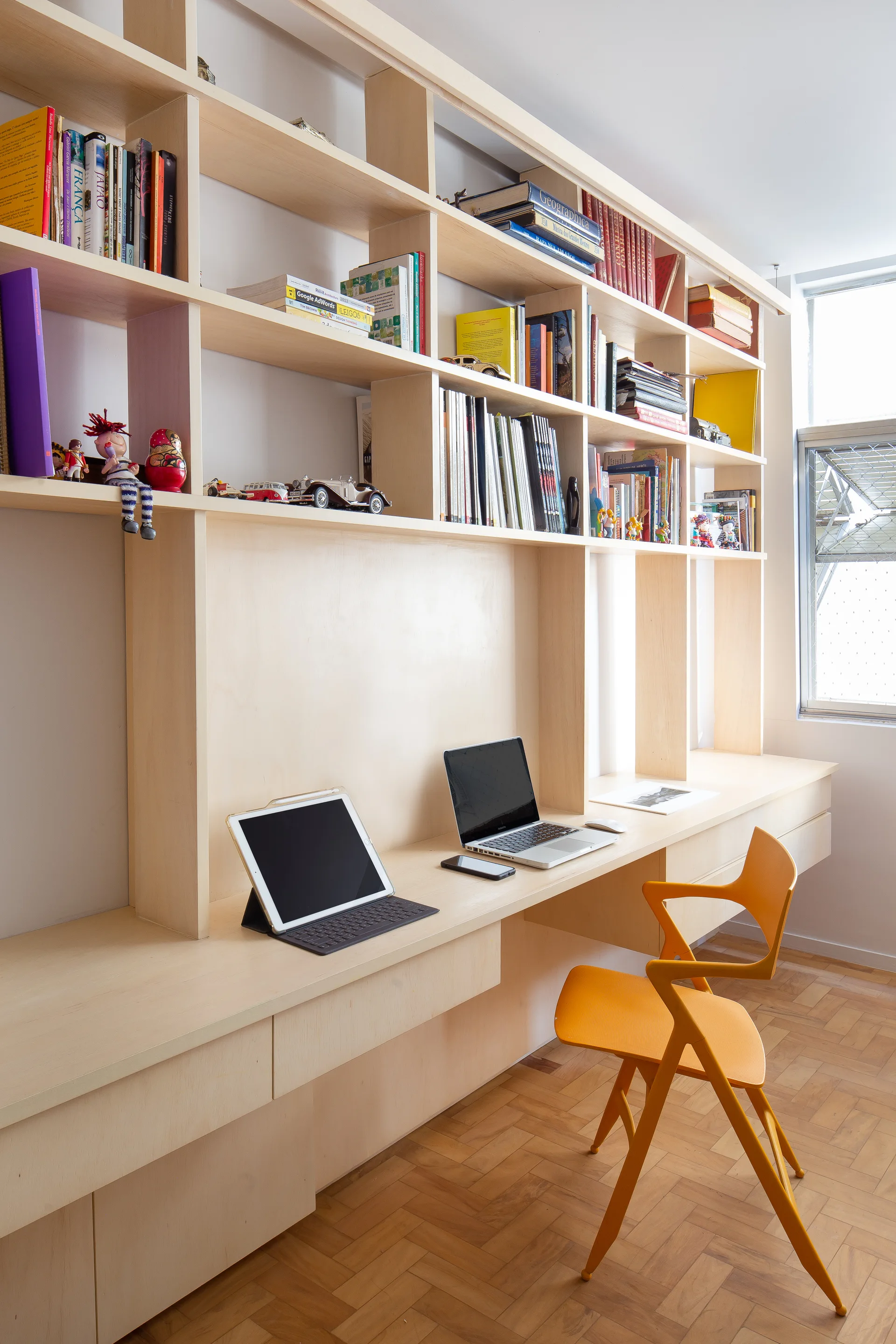 A light wood built-in workstation with bookshelves and a bright orange chair sits on herringbone parquet flooring.