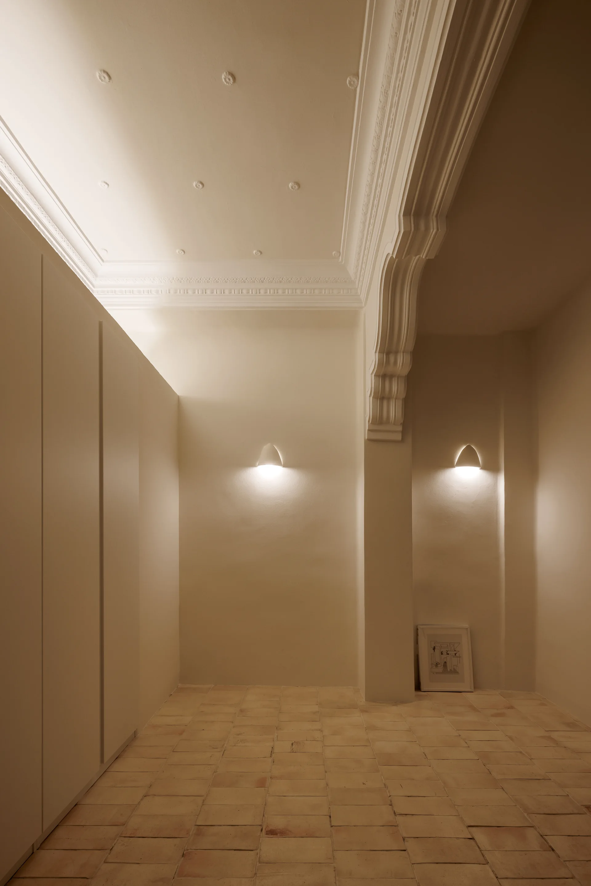 A minimalist hallway features cream walls, terracotta floor tiles, integrated flat-front wardrobes, and ornate neoclassical ceiling moldings.