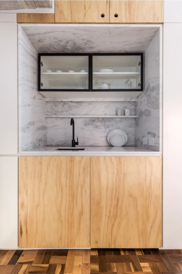 A minimalist kitchenette featuring light wood lower cabinets, a marble alcove with a black faucet, and a reeded glass upper cabinet.