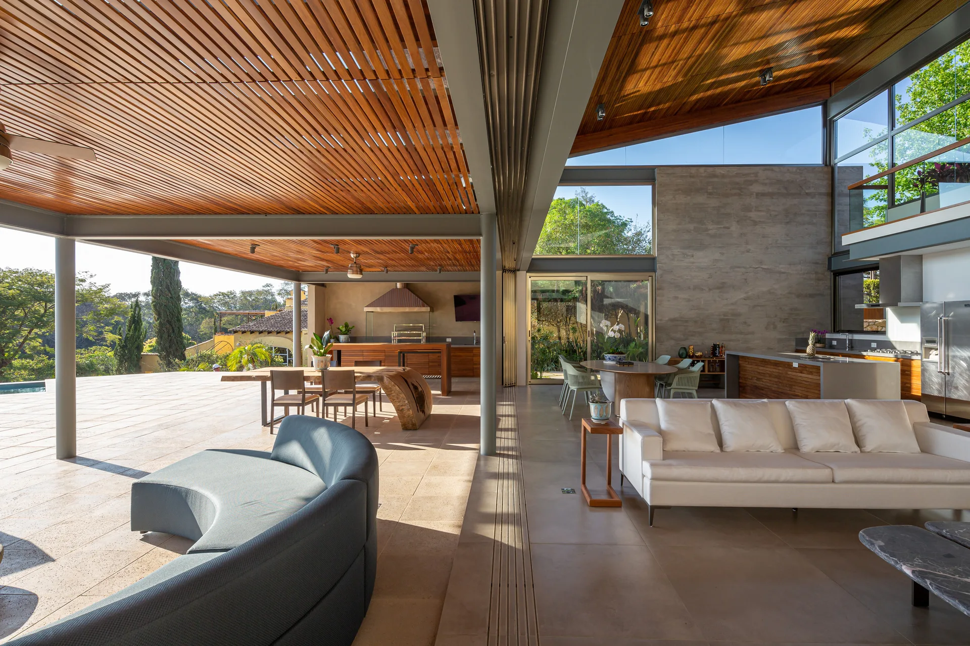 A modern open-concept living room and kitchen seamlessly connect to a covered outdoor patio under a continuous wood slatted ceiling.