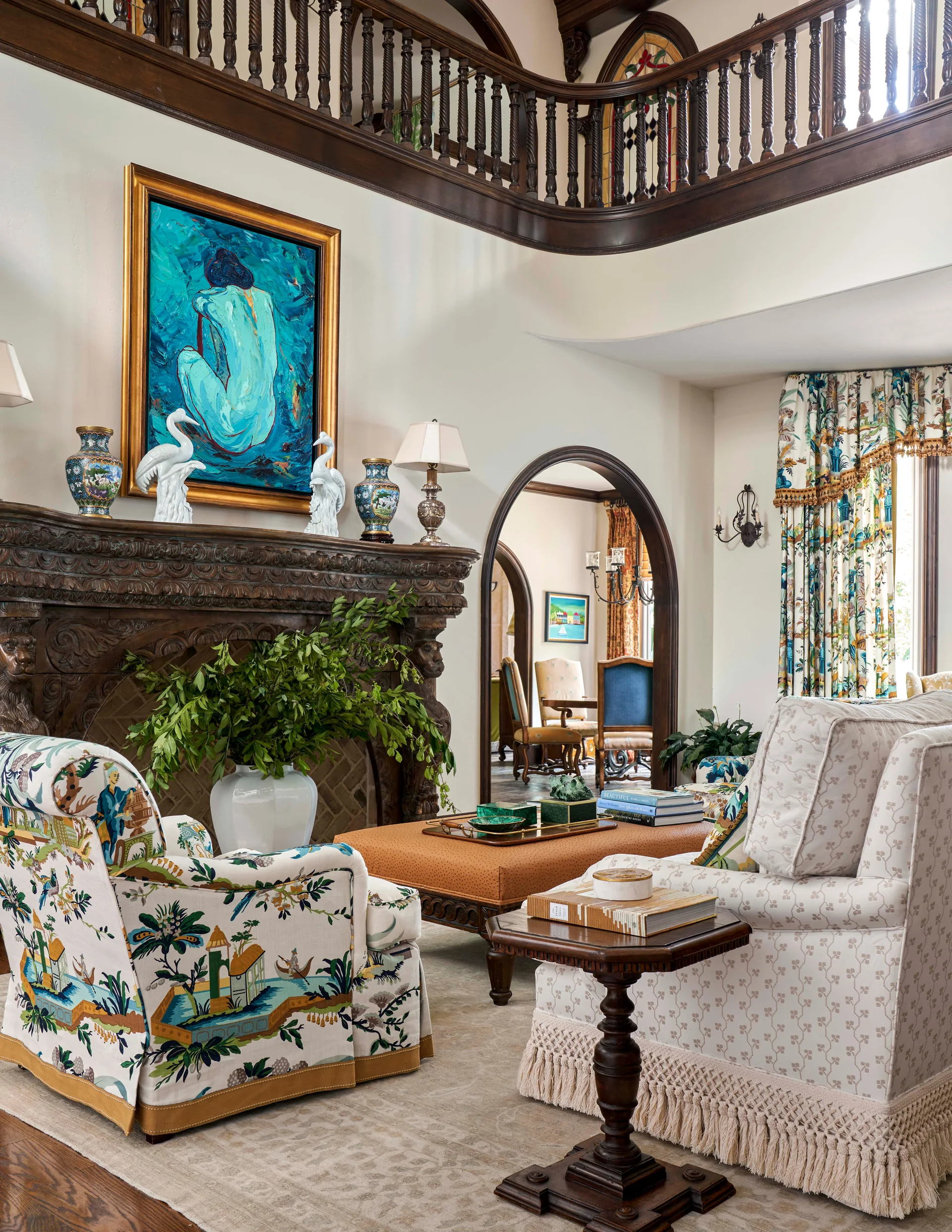 A formal living room features an ornate carved wood fireplace, a vibrant blue painting, patterned armchairs, and a central orange ottoman.
