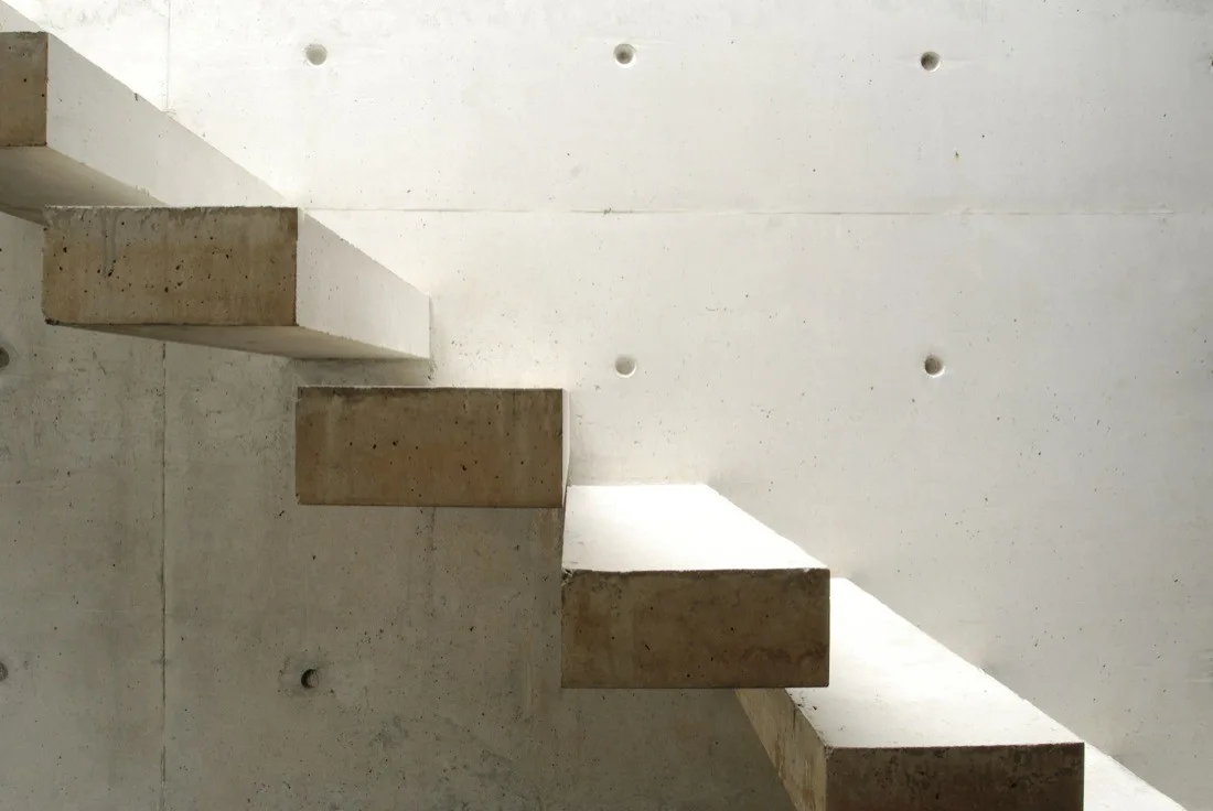 Floating concrete stair treads project from a raw, pitted off-white concrete wall in a minimalist architectural space.