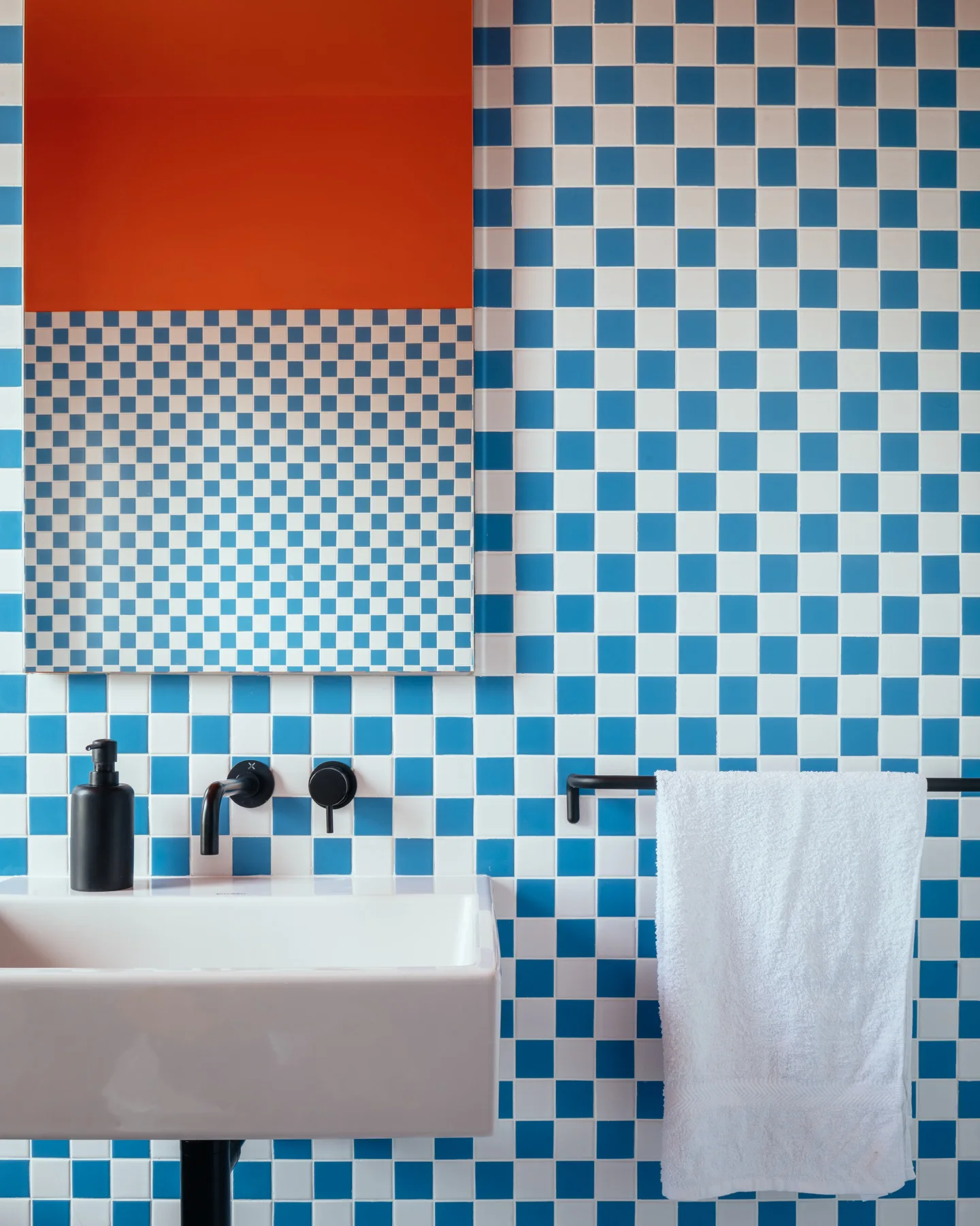 A bathroom sink area features blue and white checkered wall tile, a white wall-mounted basin, matte black faucet, and an orange accent on the mirror.