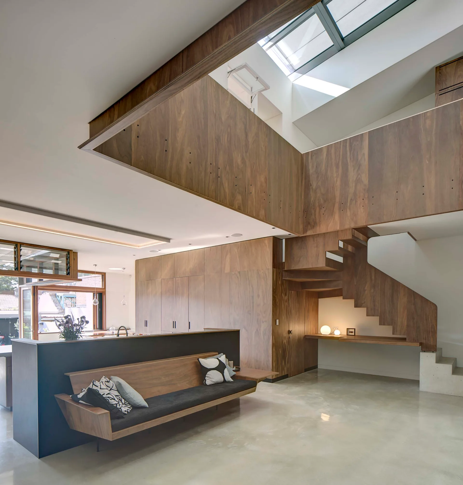 A modern open-plan interior with extensive warm wood paneling, a floating wooden staircase, polished concrete floors, and skylights overhead.