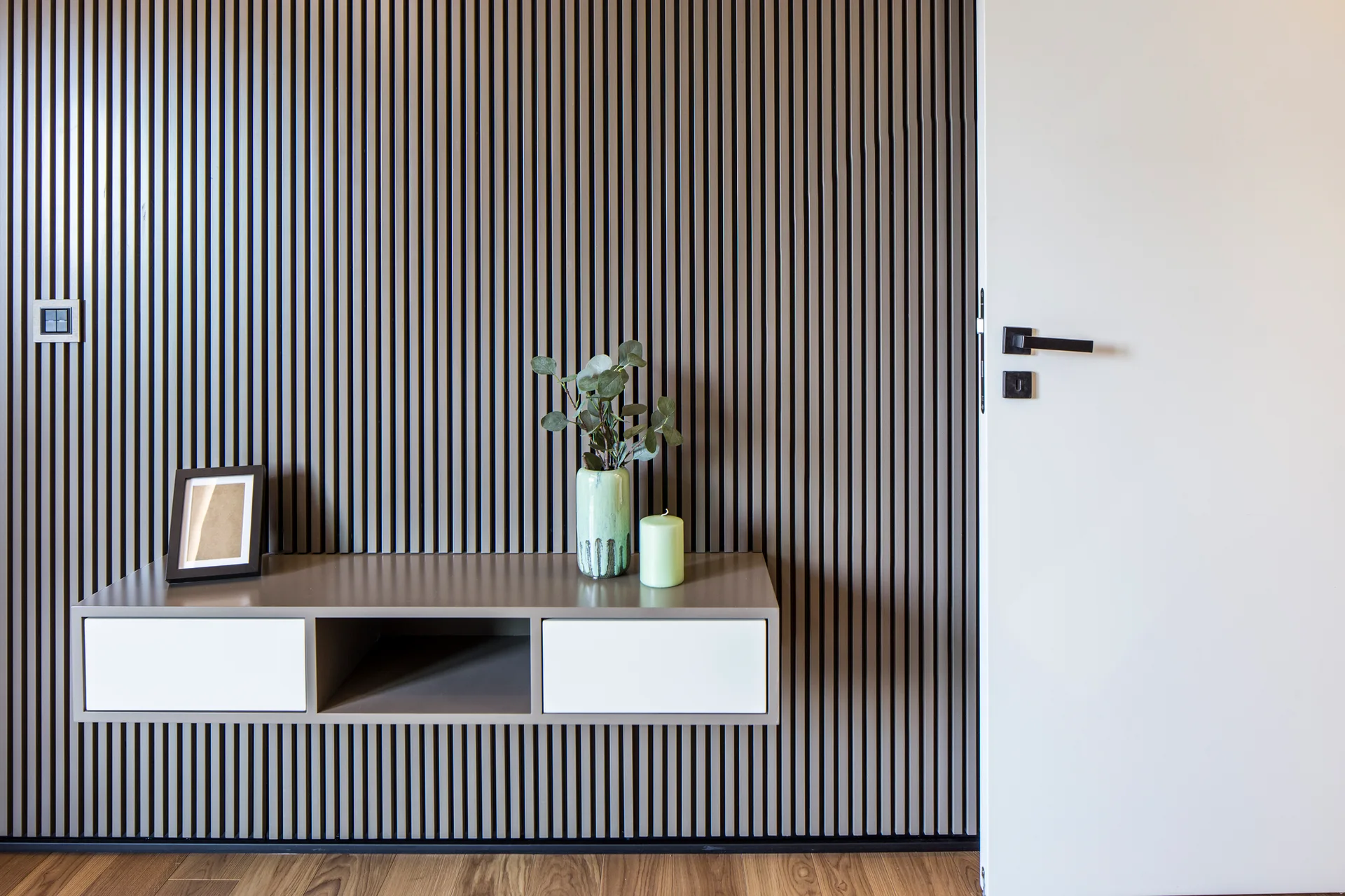 A wall covered in vertical brown wood slats features a mounted floating console with white drawers below a light green vase and a candle.