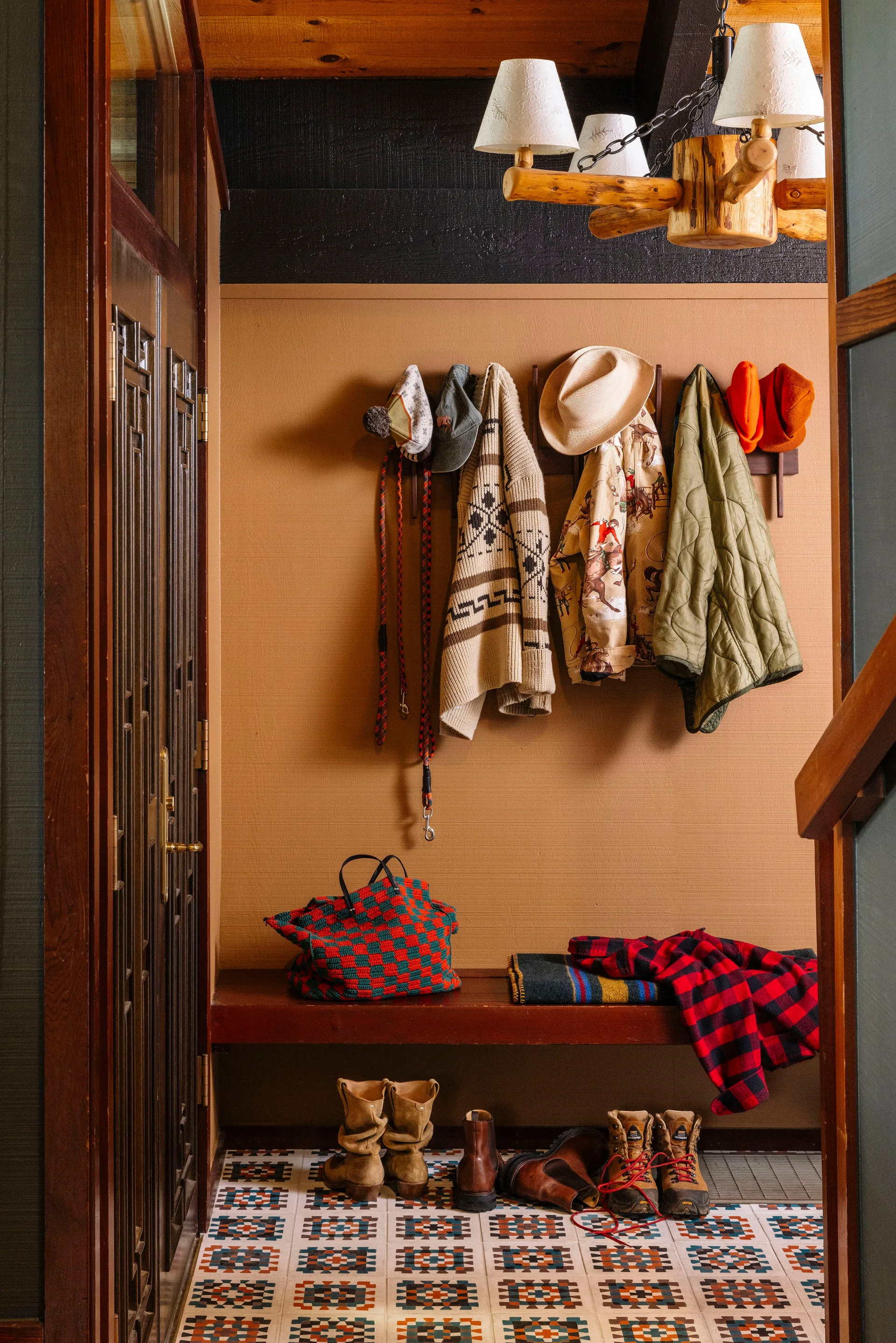 A rustic mudroom entryway features a wooden bench, a coat rack with hanging jackets, patterned floor tiles, and a log chandelier.