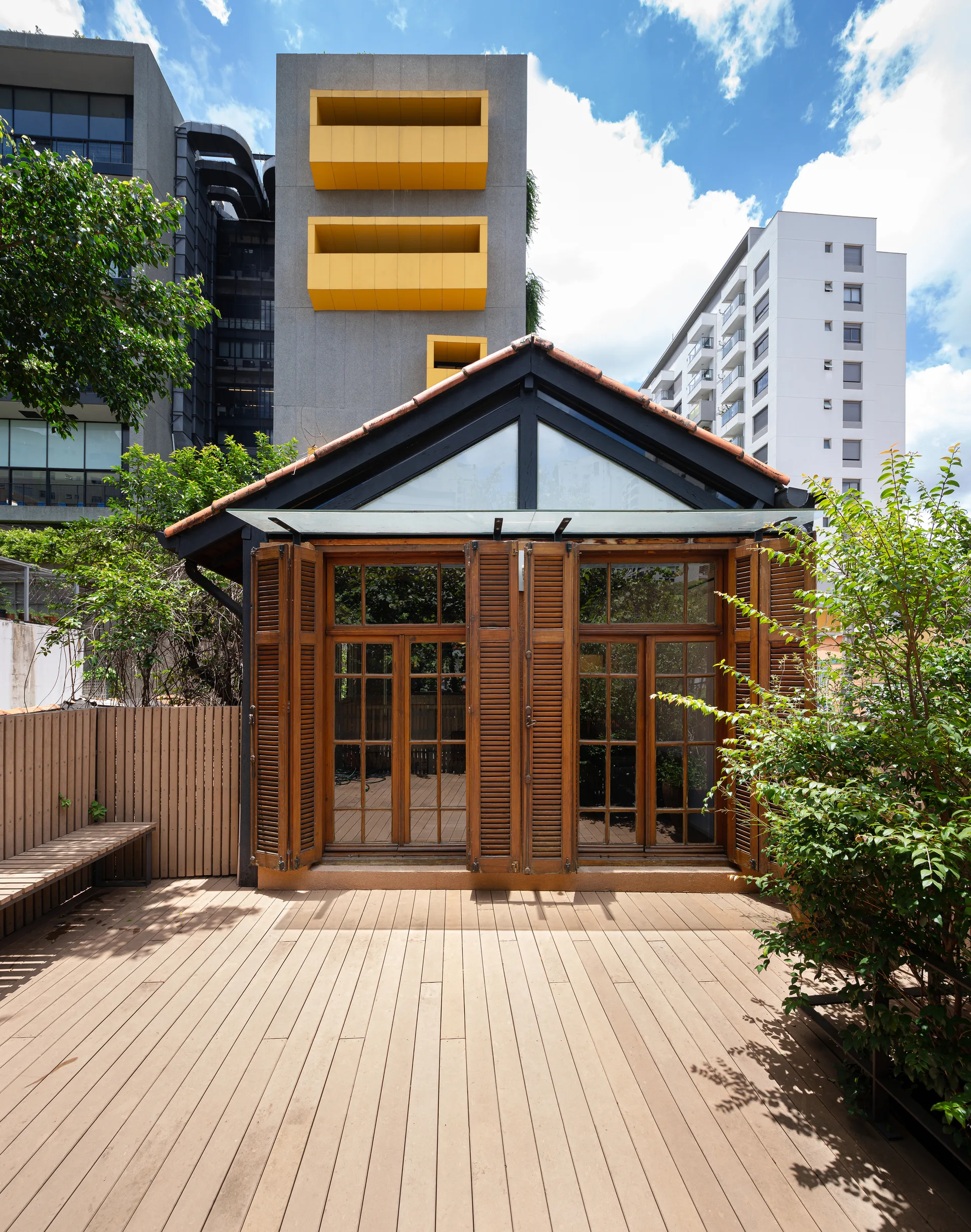 An outdoor wooden rooftop terrace features a small gabled building with warm brown folding shutters set against modern high-rise buildings.