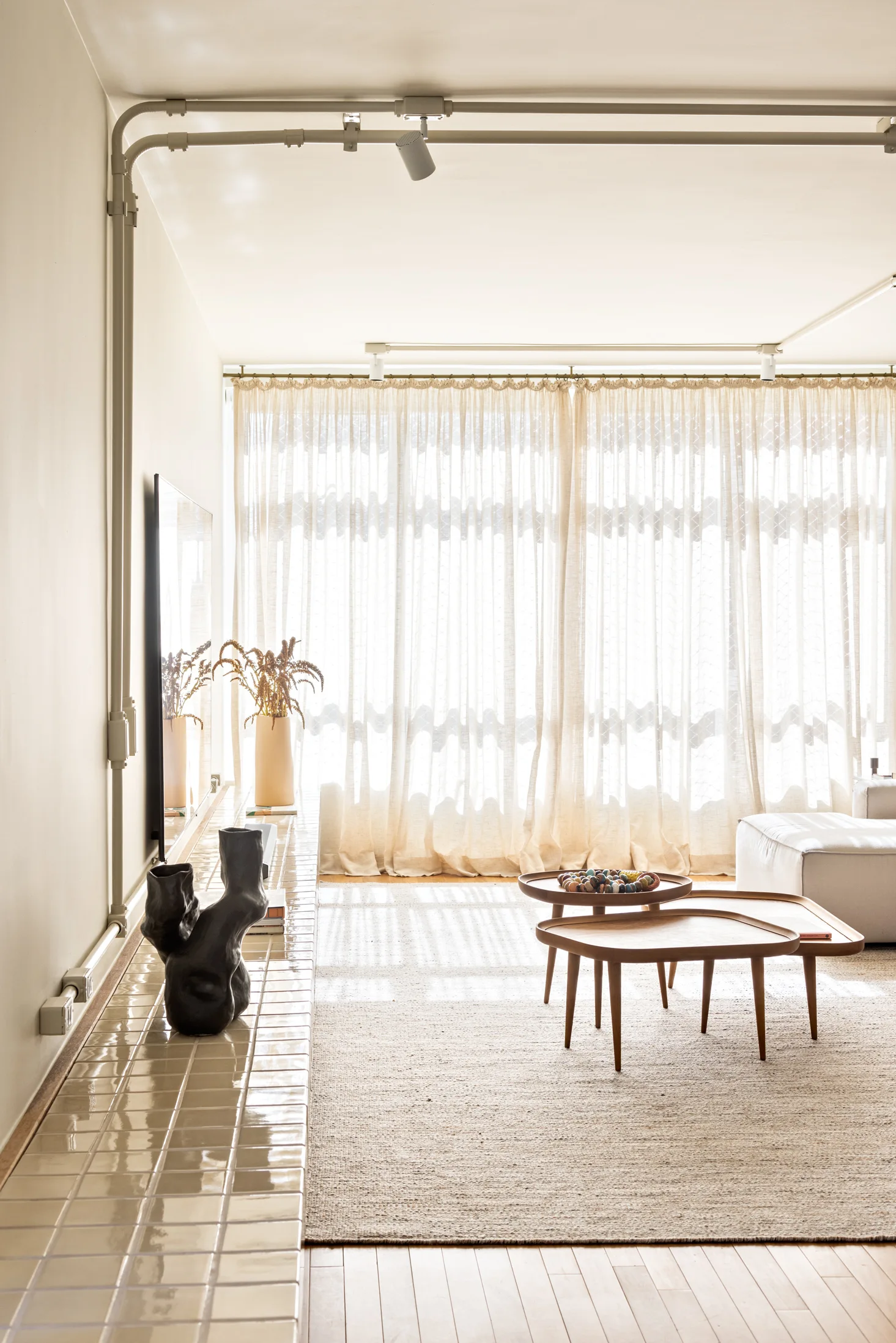 A bright living room features a long glossy tiled bench, nested wood coffee tables on a textured rug, and sheer curtains filtering natural light.