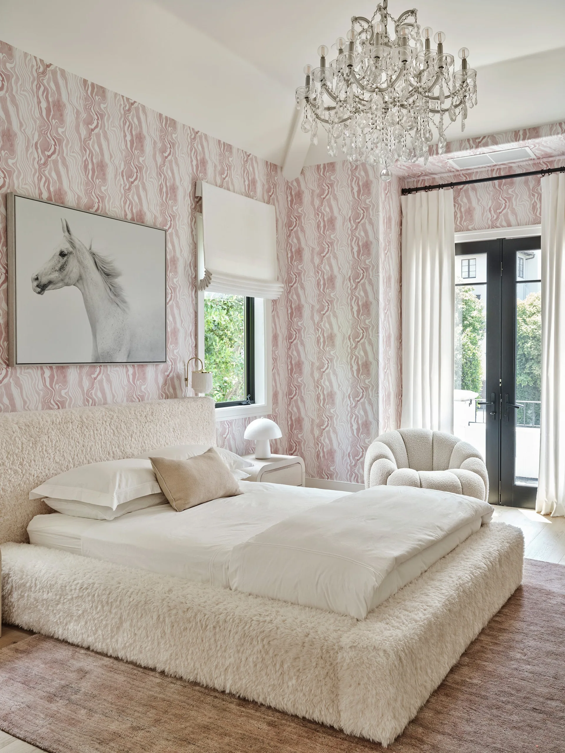 A contemporary bedroom features pink wavy-patterned wallpaper, a plush cream bed frame, and an ornate crystal chandelier.