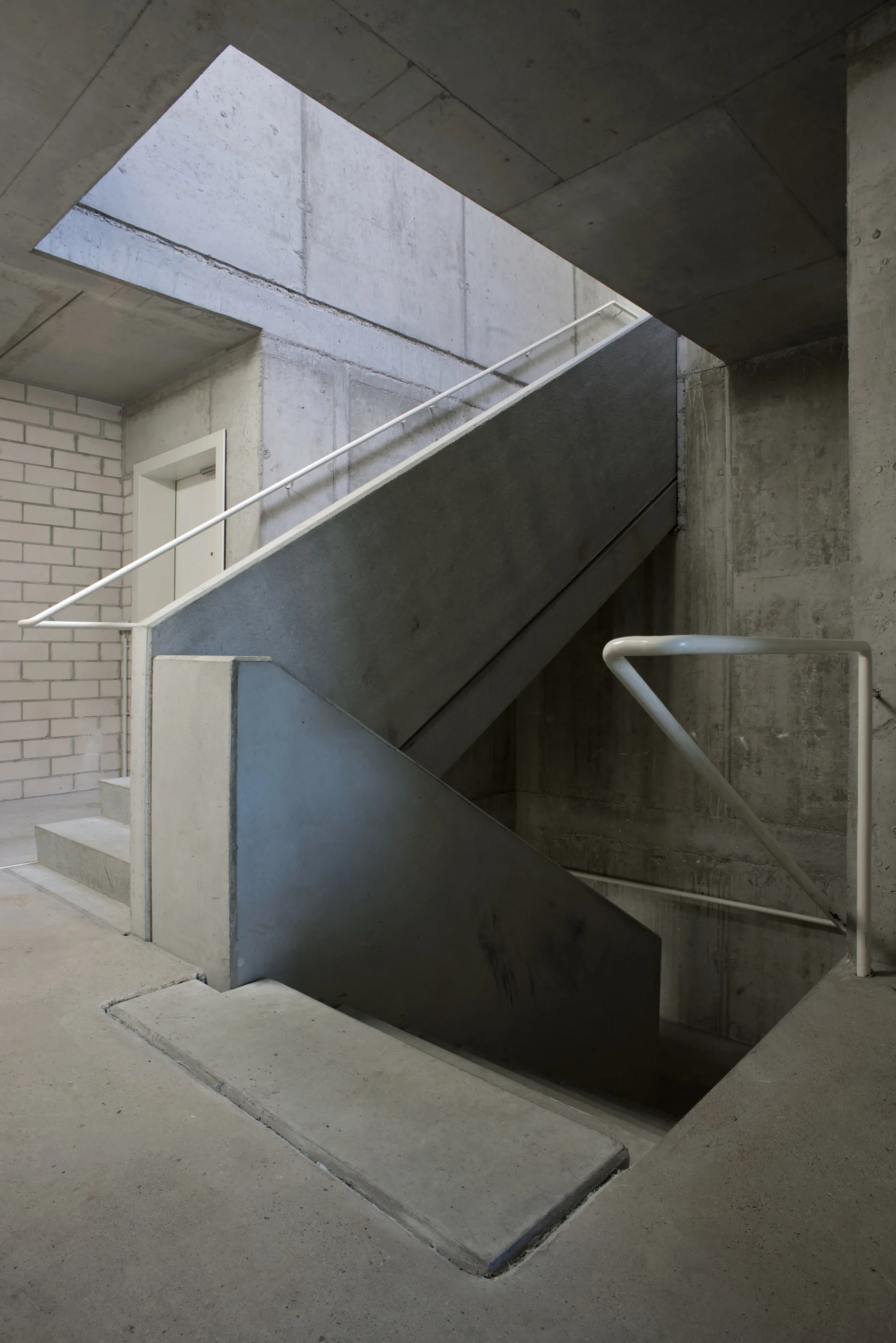 A brutalist concrete staircase with raw grey finishes features slim white metal railings beneath a rectangular ceiling skylight.