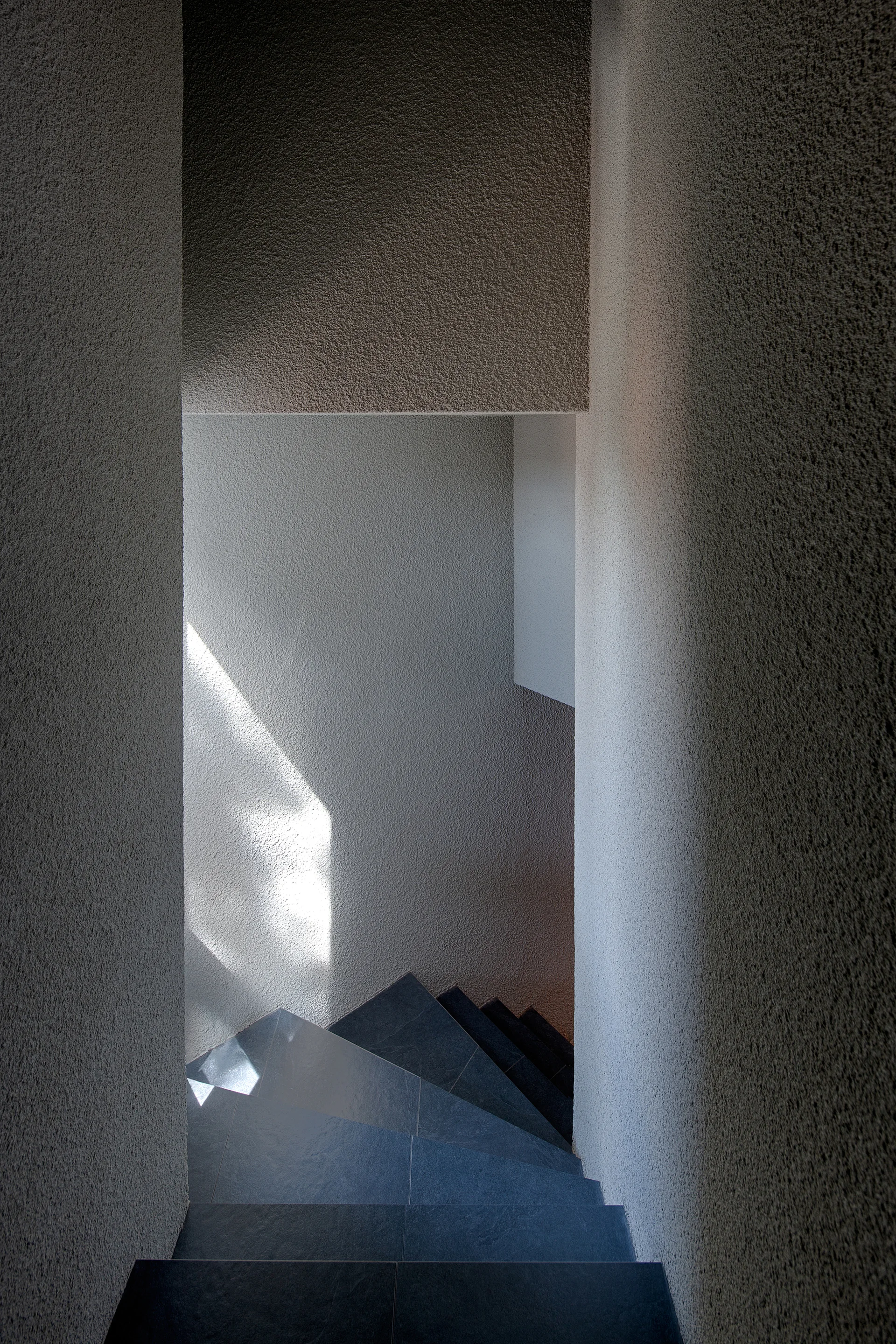 A minimalist stairwell features heavily textured grey plaster walls and dark stone steps winding downwards with sunlight casting sharp shadows.