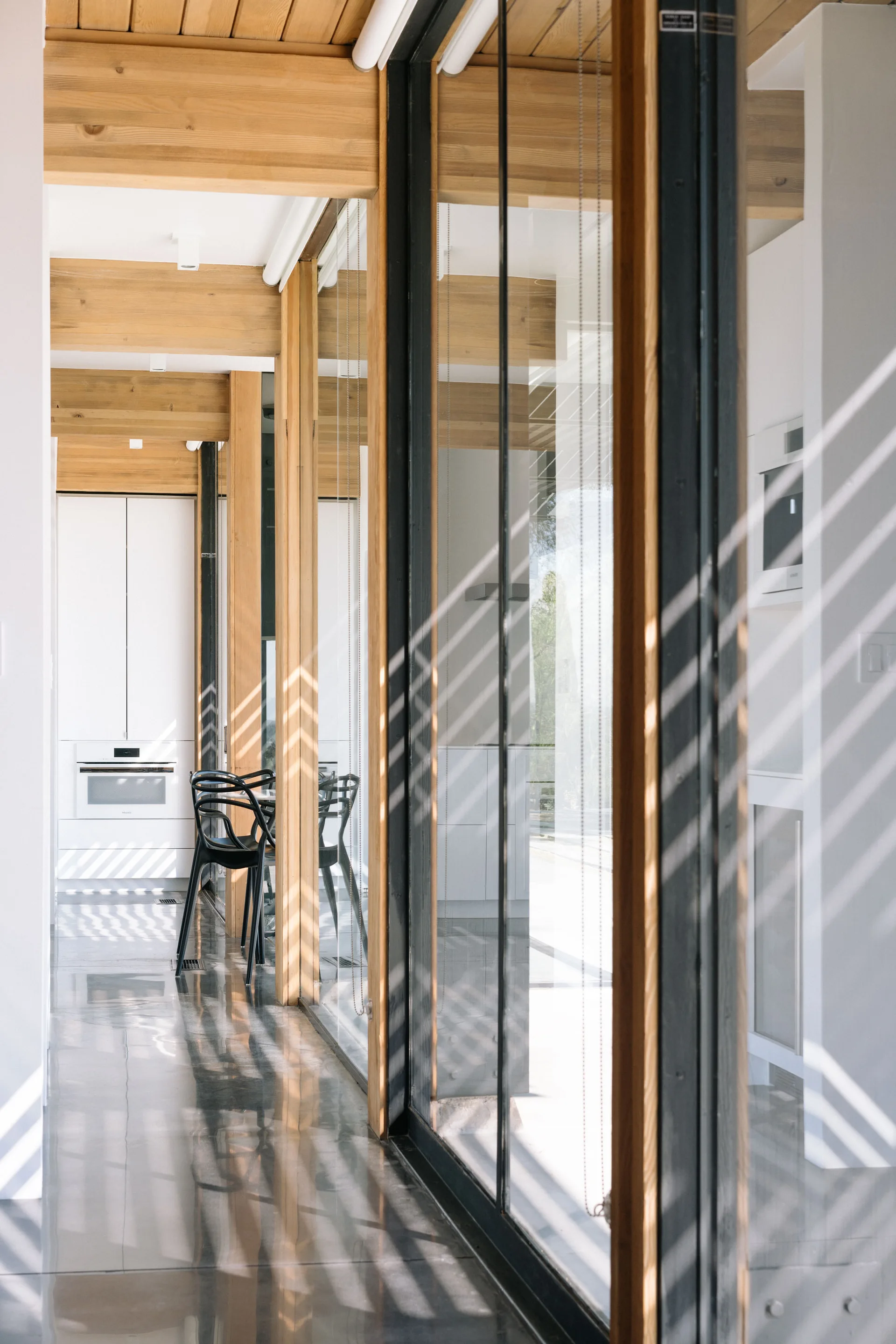 A modern kitchen corridor features polished grey floors, exposed wooden ceiling beams, white cabinetry, and a floor-to-ceiling glass wall.