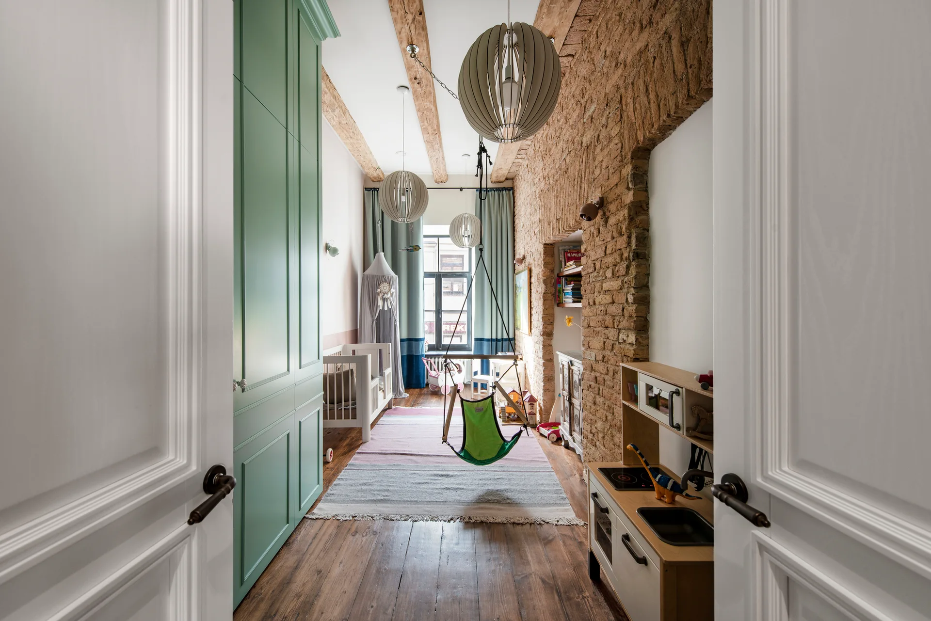 A long kids' room featuring an exposed brick wall, timber ceiling beams, a sage green wardrobe, and a swing chair suspended from the ceiling.