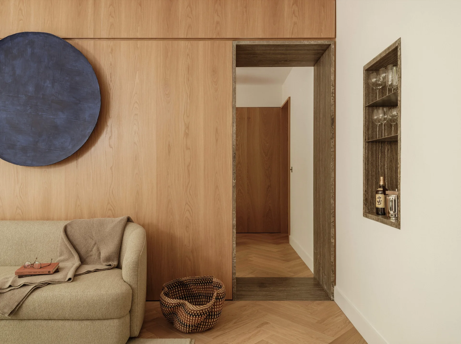 A beige sofa rests near a wood-paneled wall, featuring an open doorway and a recessed stone-lined display niche with glassware on the adjacent wall.