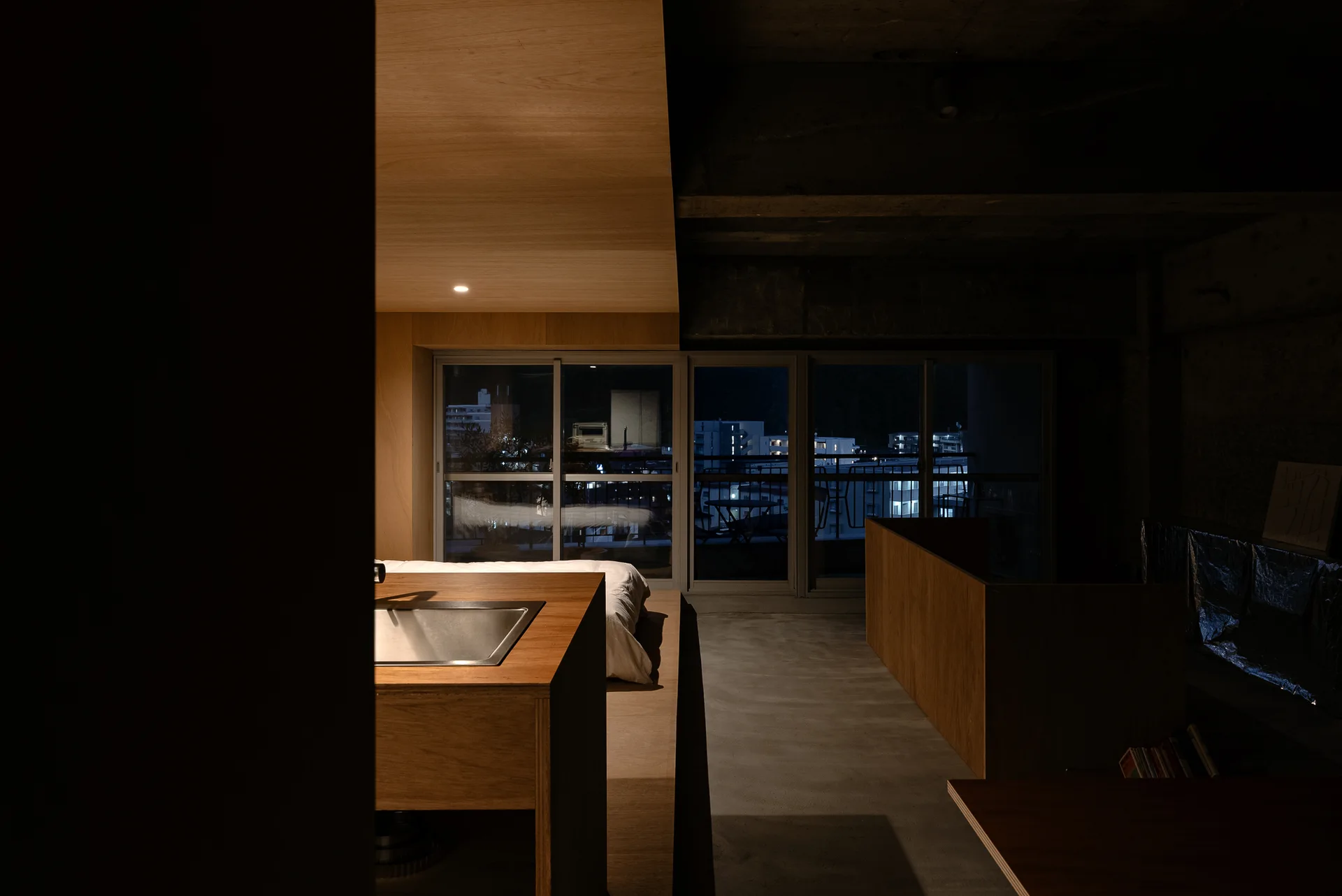 A minimalist studio features warm wood paneling, an integrated kitchen island, concrete surfaces, and sliding glass doors showing a city skyline at night.