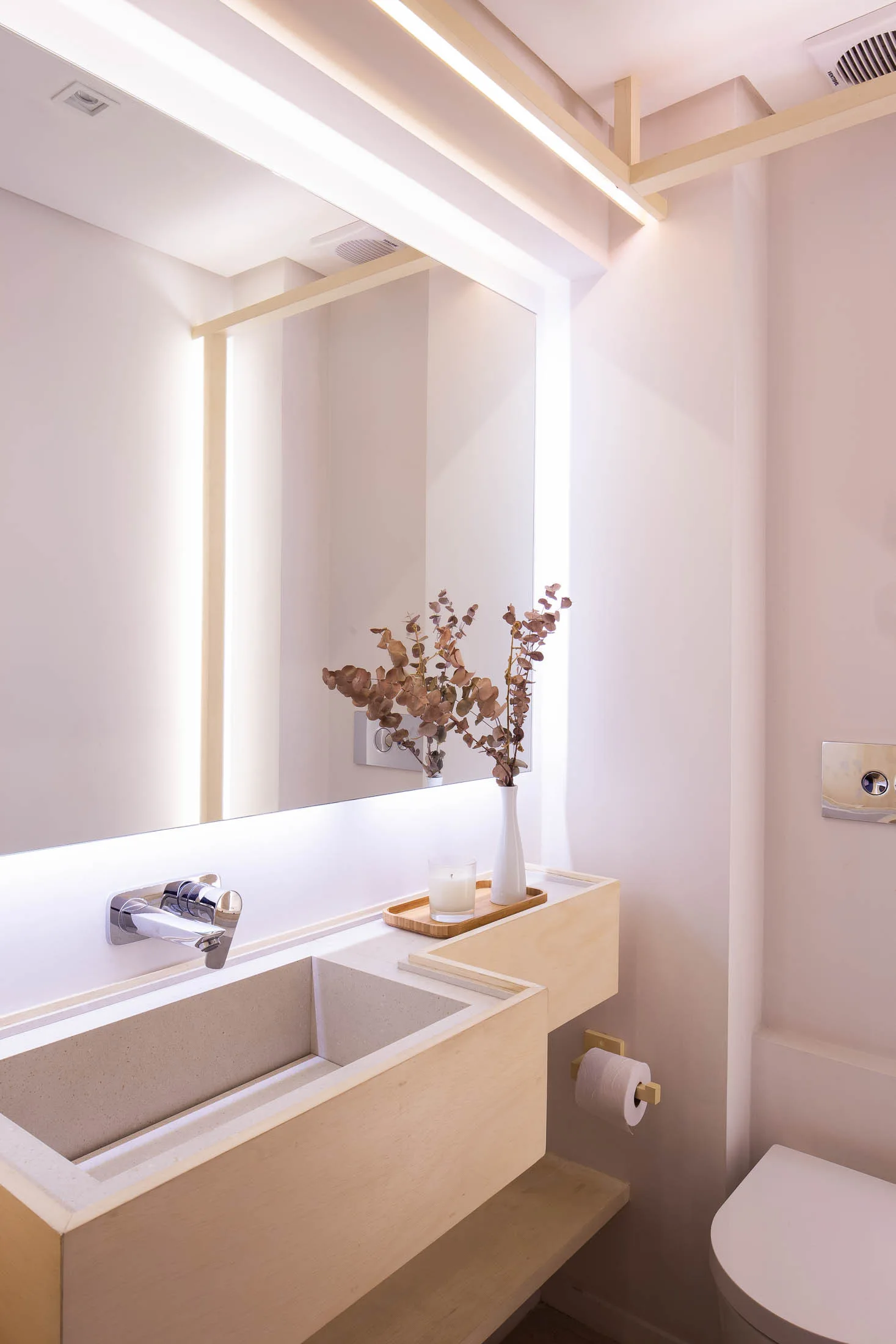 A minimalist bathroom features a light wood vanity with an integrated stone basin, a wall-mounted faucet, and a large backlit mirror.