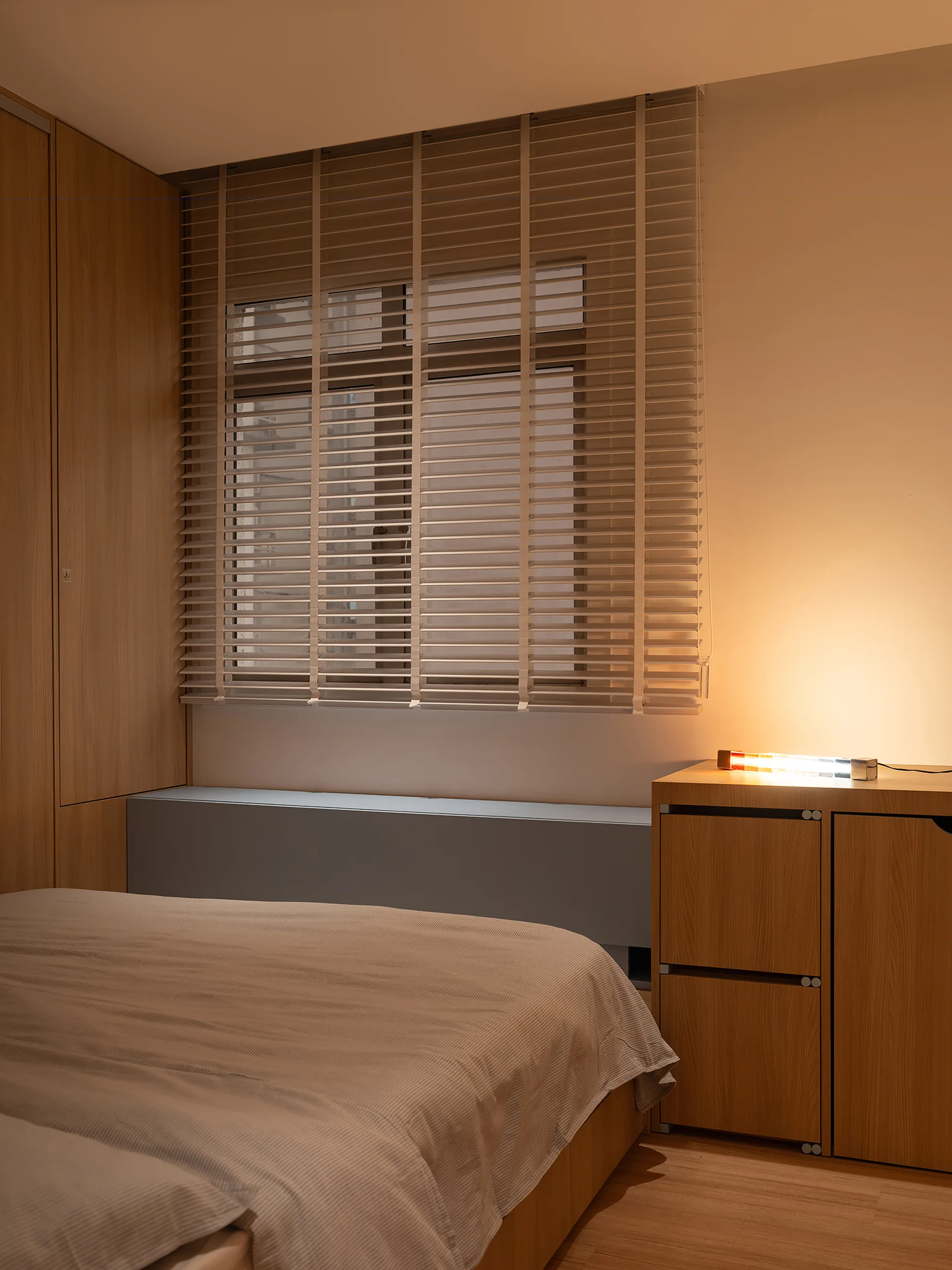 A modern bedroom features a low bed with striped bedding, a wood wardrobe, a storage chest under horizontal blinds, and a glowing table lamp.
