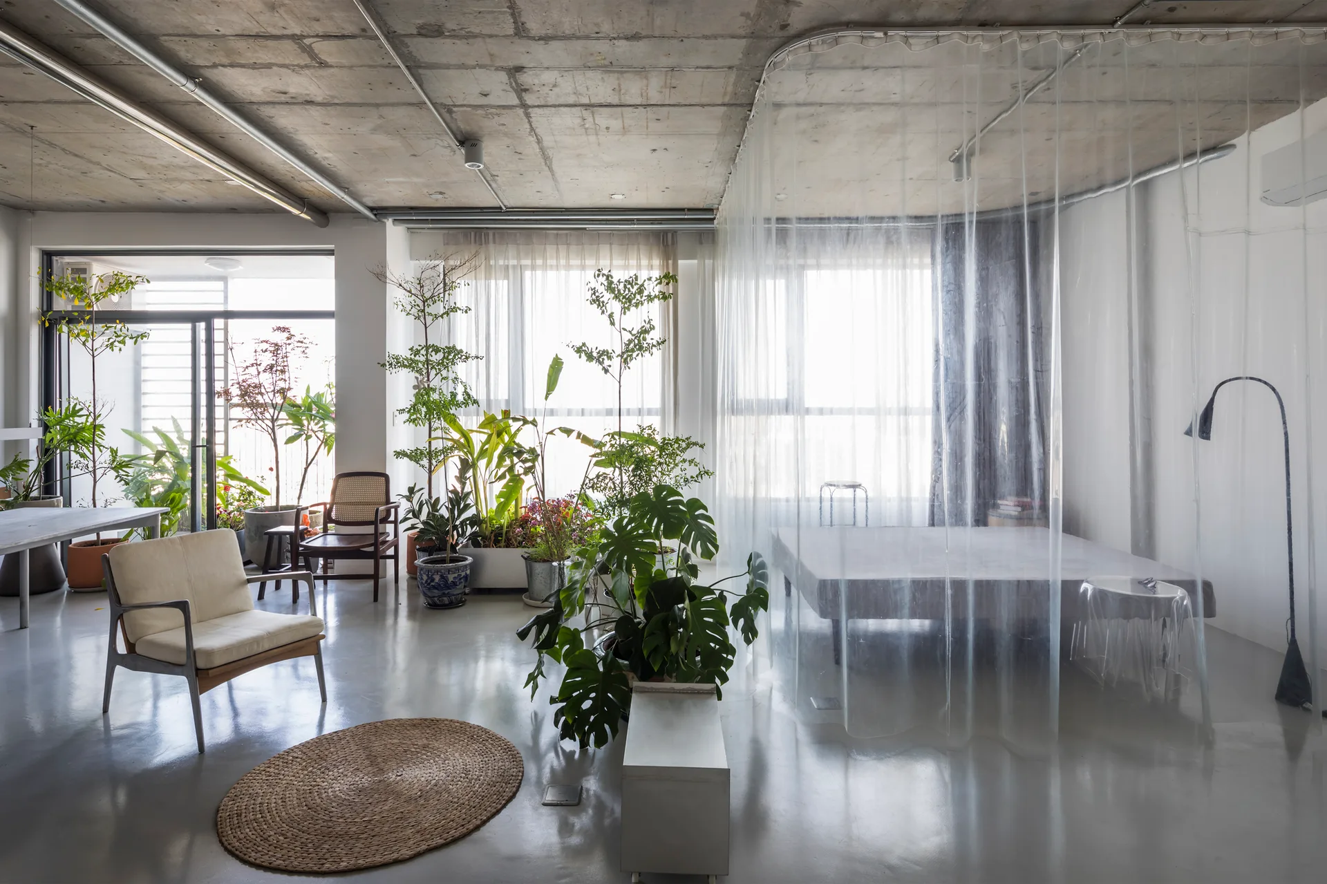 An industrial loft bedroom features an exposed concrete ceiling, a polished grey floor, indoor plants, and a bed enclosed by a clear plastic curtain.