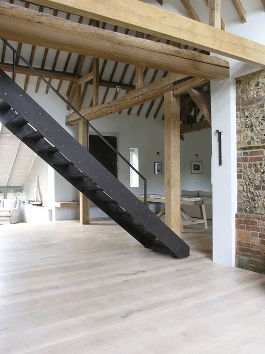 A modern black metal staircase ascends beneath heavy exposed timber ceiling beams in a room with light wood flooring and white walls.
