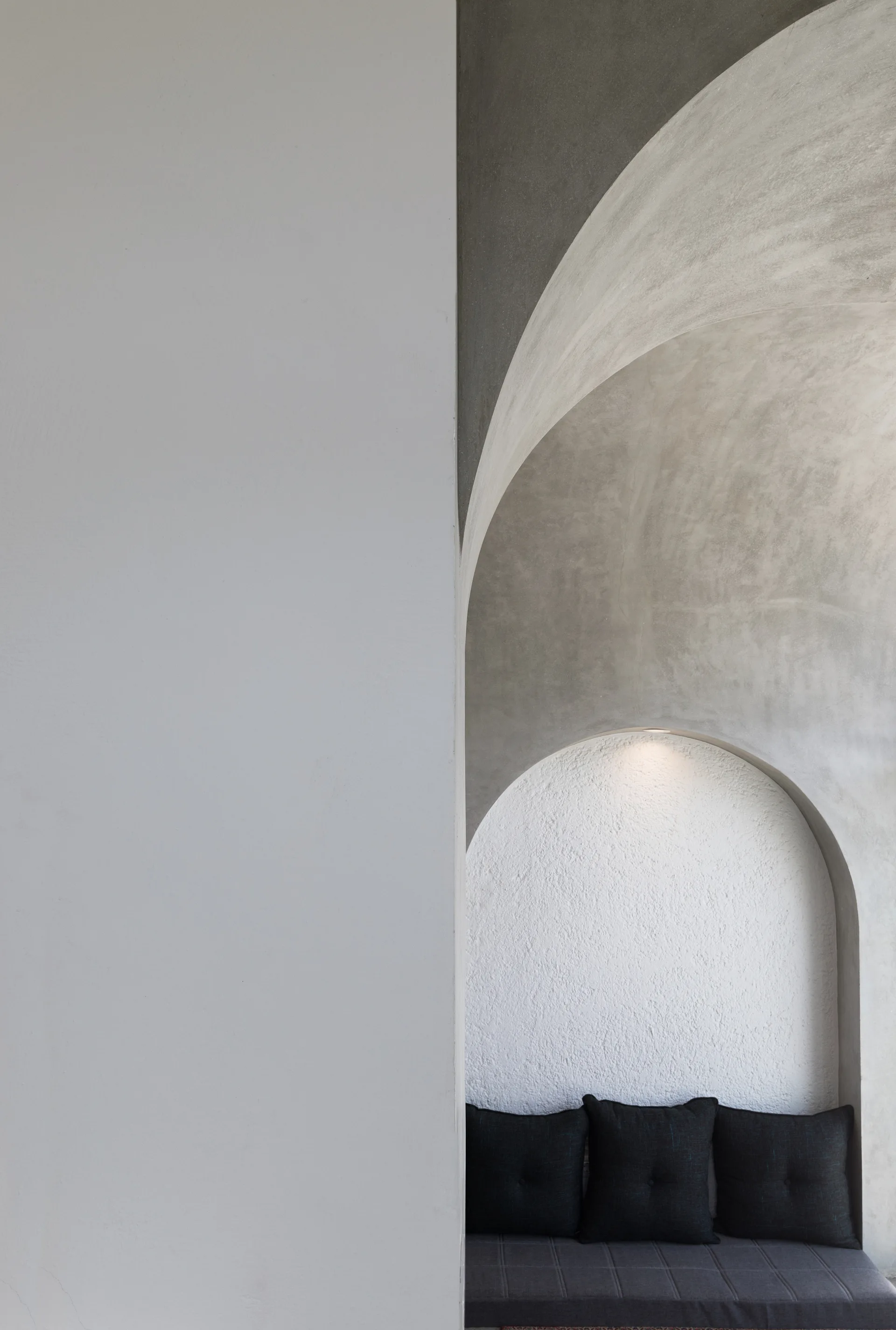 A minimalist architectural space featuring a textured plaster wall niche with a built-in dark grey bench and a curved archway overhead.
