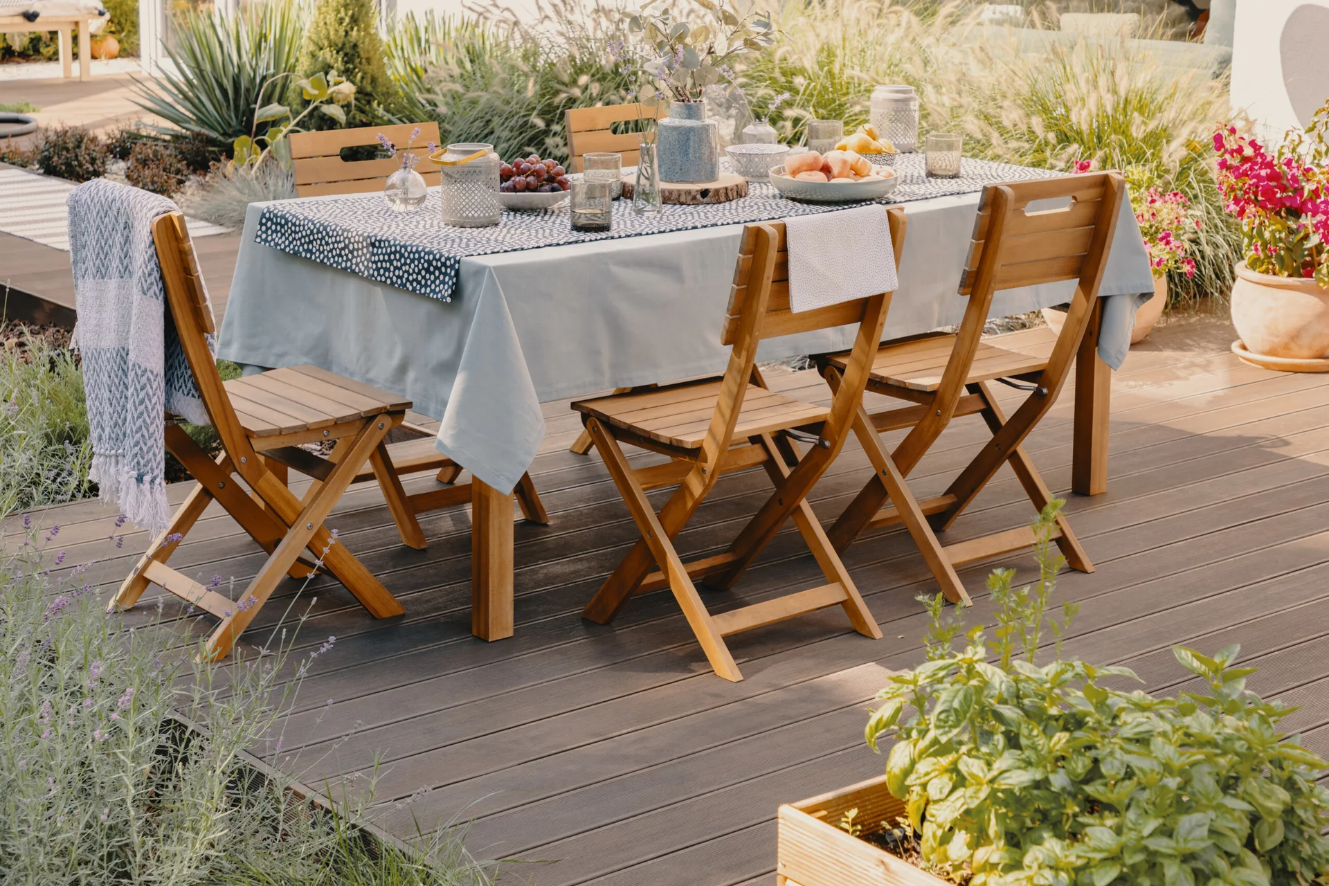 A wooden dining table with a light blue tablecloth and matching folding chairs sits on a dark composite deck surrounded by garden plants.