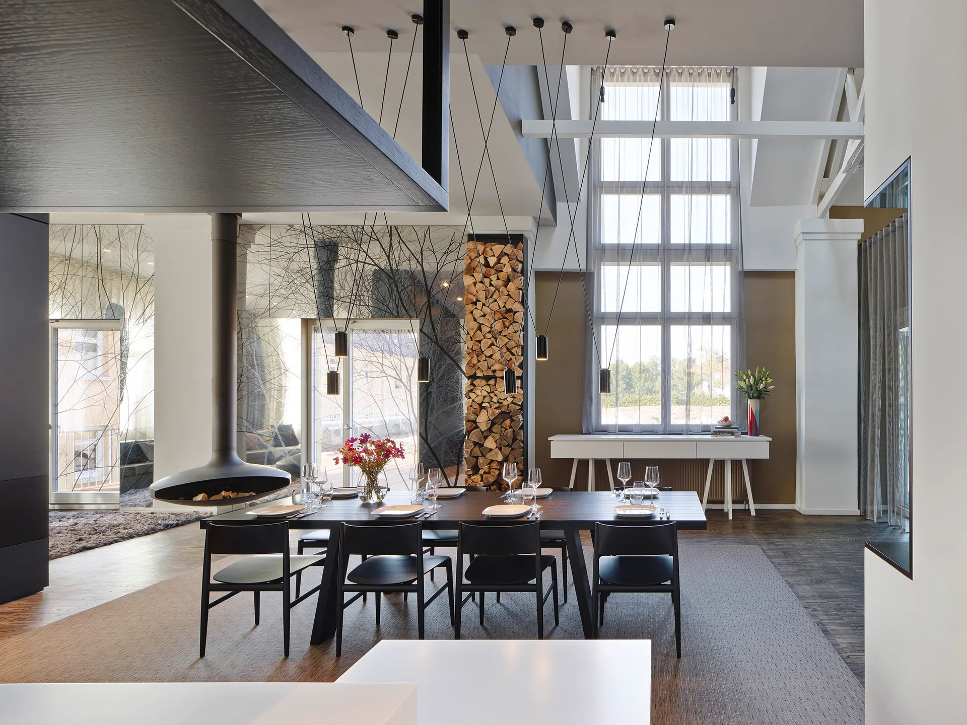 A modern dining room featuring a black table and chairs beneath slim pendant lights, next to a suspended fireplace and a vertical column of stacked firewood.