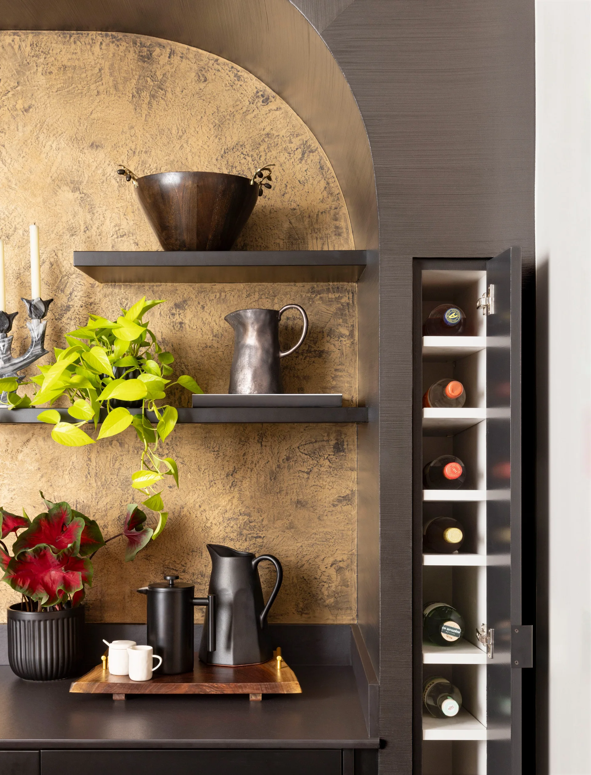 An arched built-in shelving unit with black shelves, a textured gold backing, and a narrow integrated wine cabinet holding bottles on shelves.