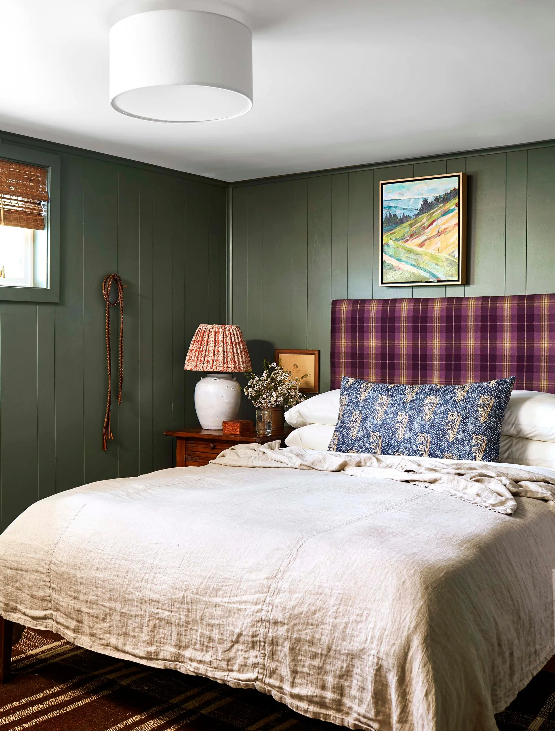 A bedroom with dark sage green wood-paneled walls, a bed featuring a purple plaid headboard, neutral linen bedding, and a wooden nightstand.