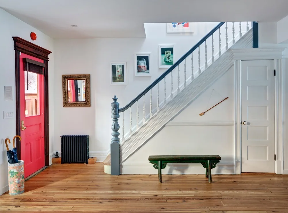 A bright entryway features a vibrant pink front door, a white wooden staircase with a grey handrail, and a low green bench on hardwood floors.