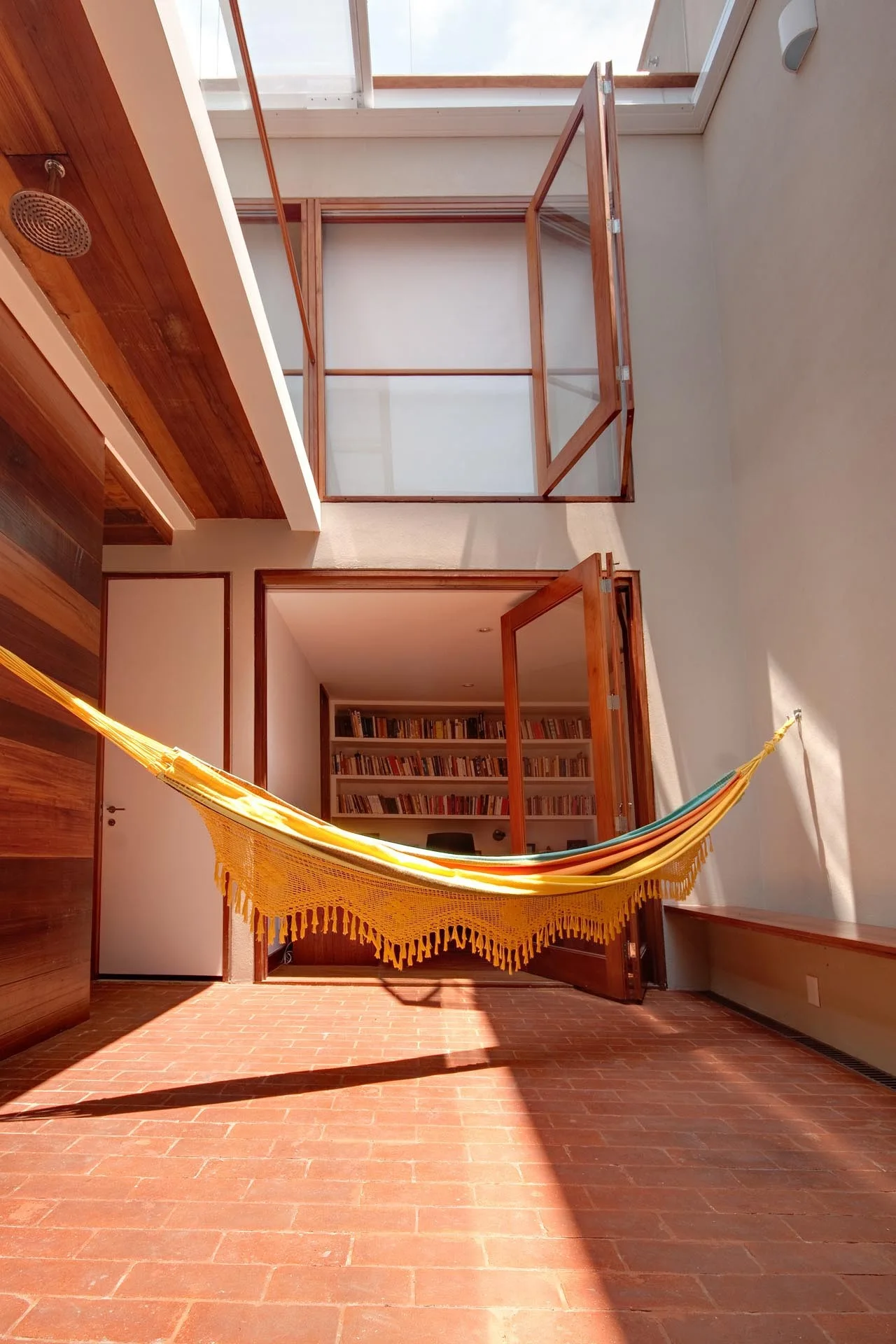 A yellow fringed hammock hangs across a sunlit courtyard with terracotta brick flooring, wooden glass doors, and a built-in bookshelf visible inside.